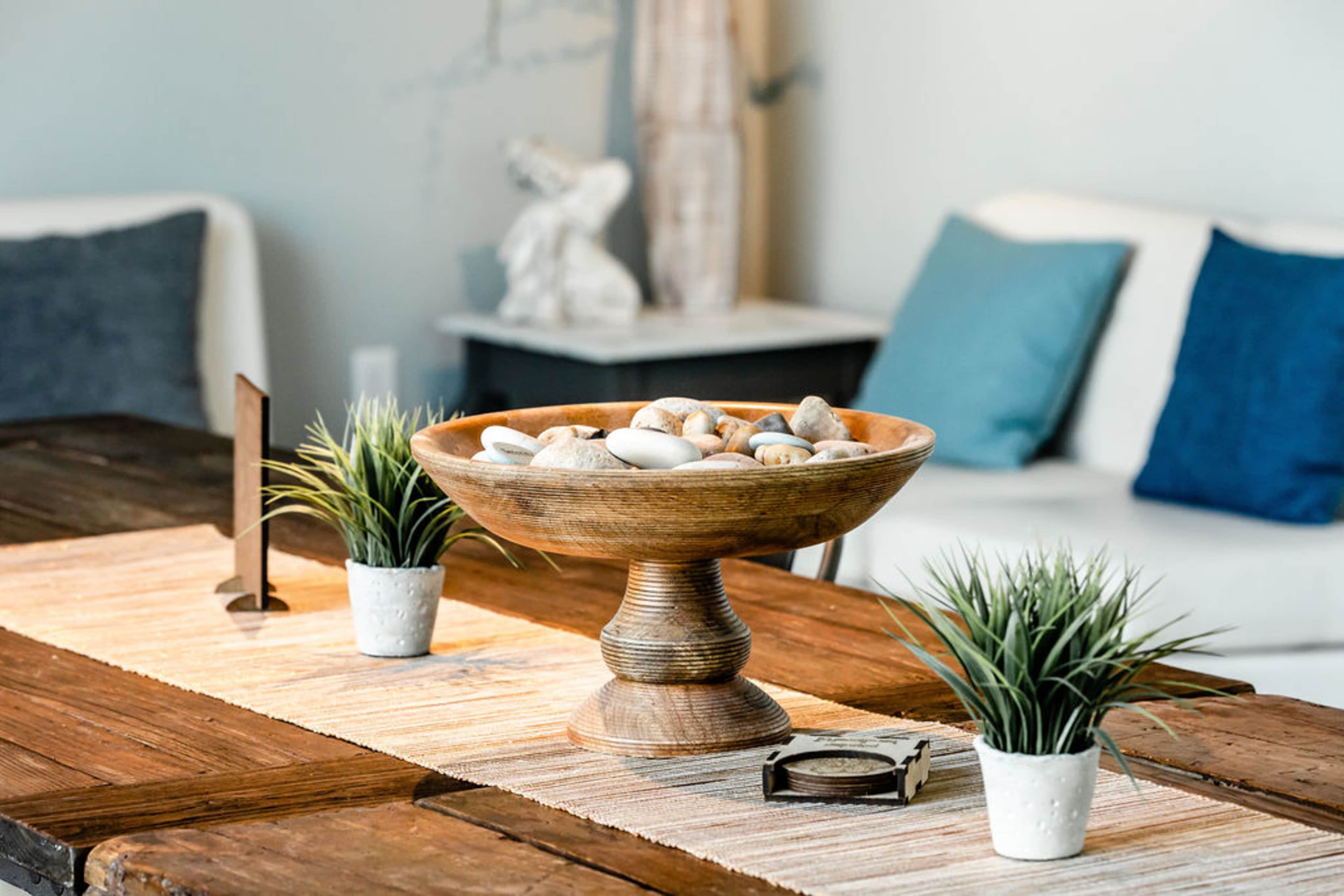 A wooden bowl filled with decorative stones sits on a table decorated with a woven table runner, flanked by small potted plants and a compass.