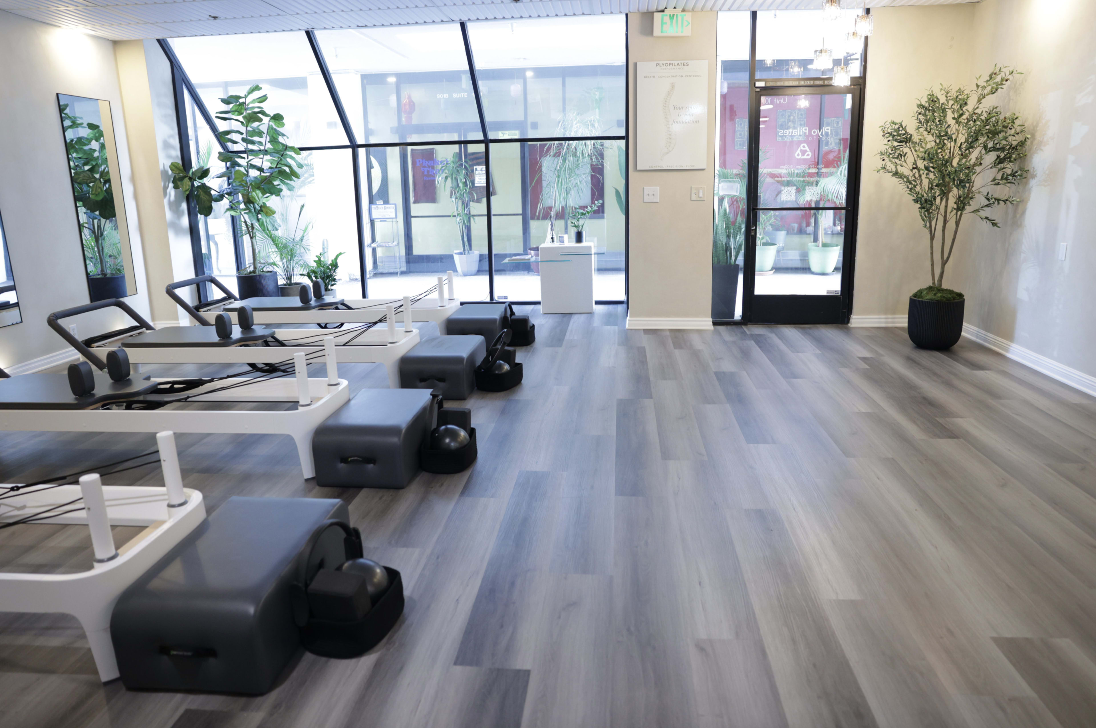 The image shows a fitness studio with reformer Pilates machines arranged neatly along one side and large windows allowing natural light to enter.