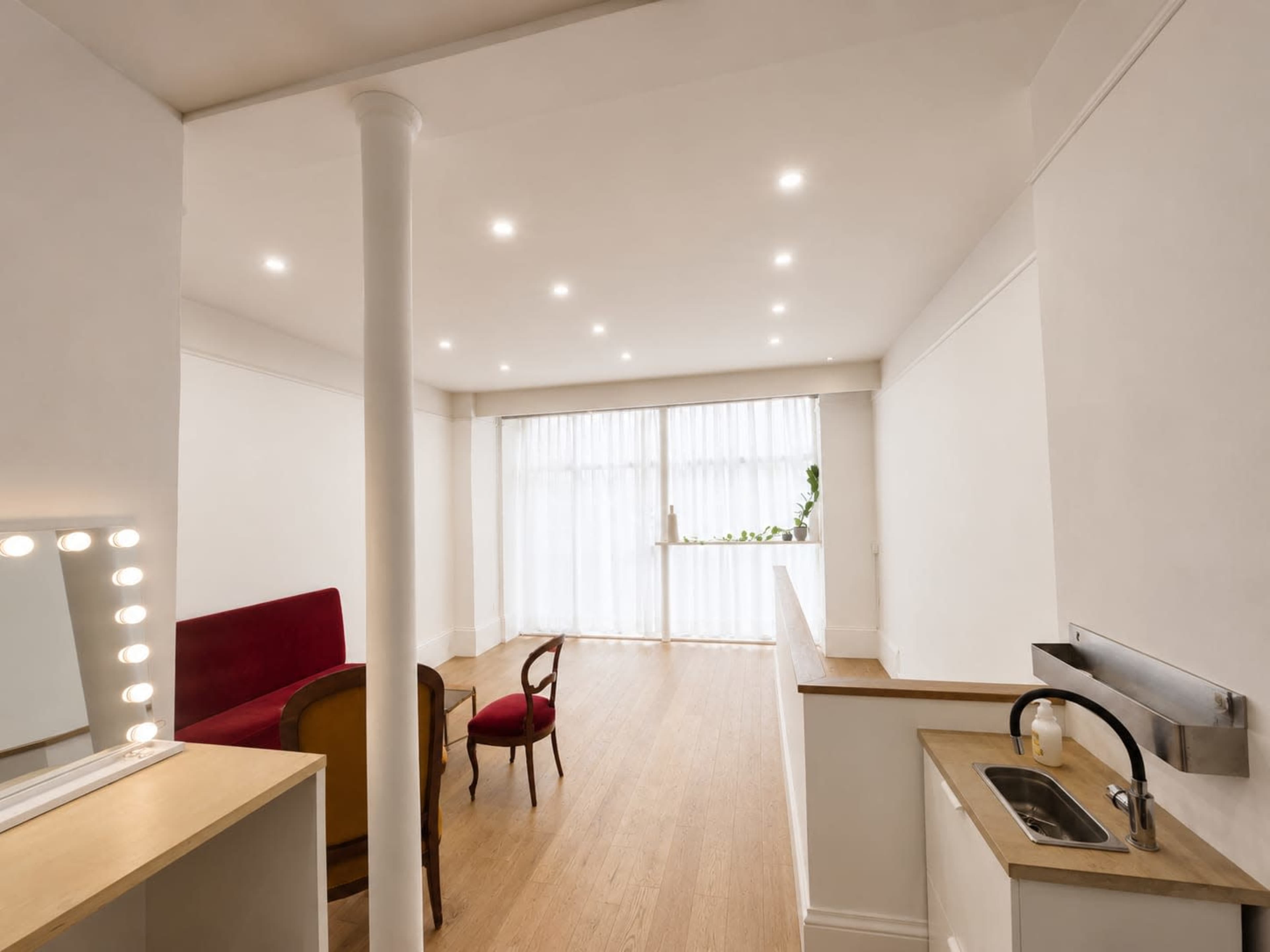 The image shows a minimalistic interior space with wooden flooring, bright lighting, a red sofa, a small kitchen sink, and a table with chairs.
