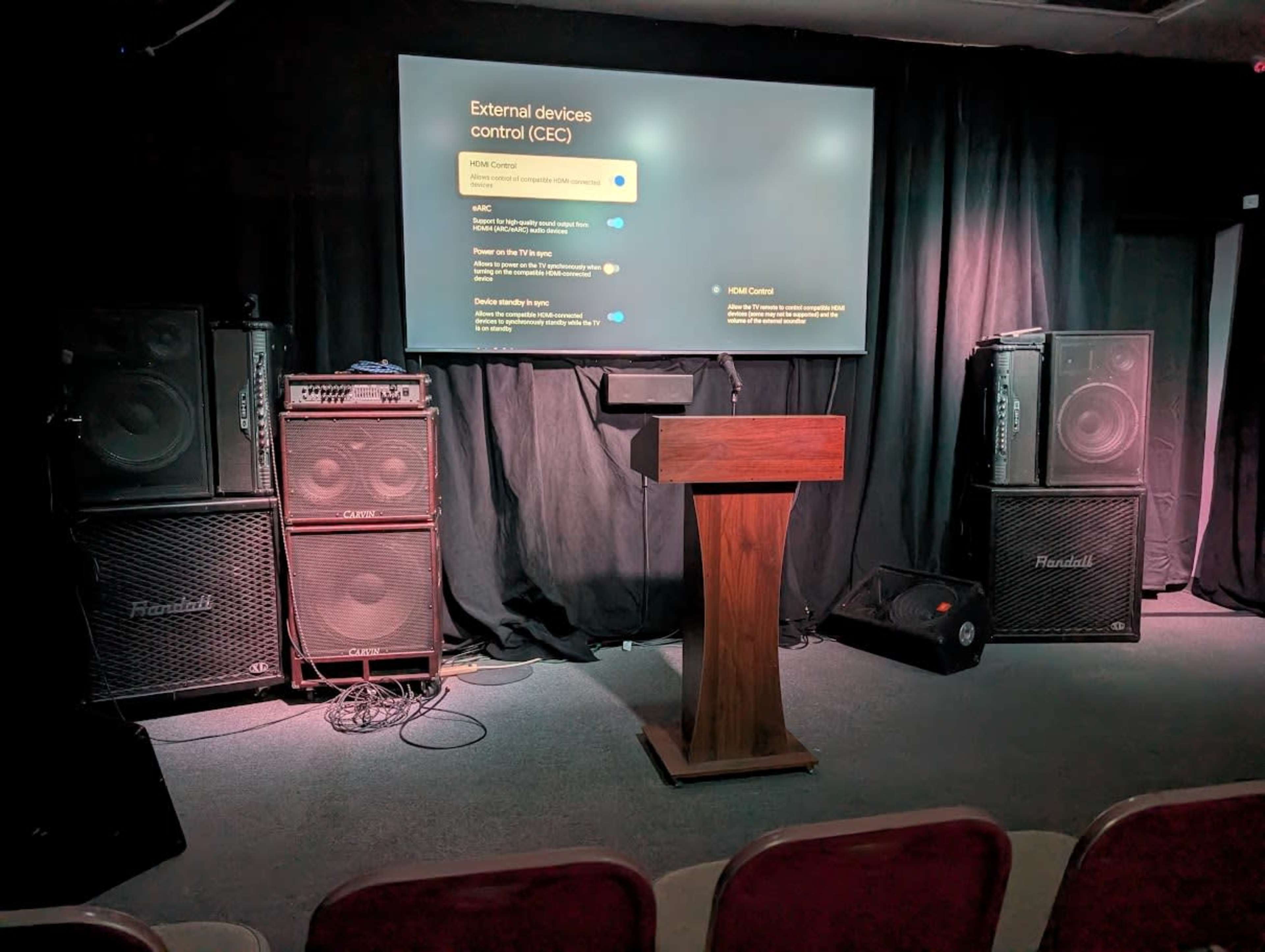 The image shows a small stage setup with a podium in the center, flanked by two large audio speakers, and a screen displaying settings for external devices in a darkened room.