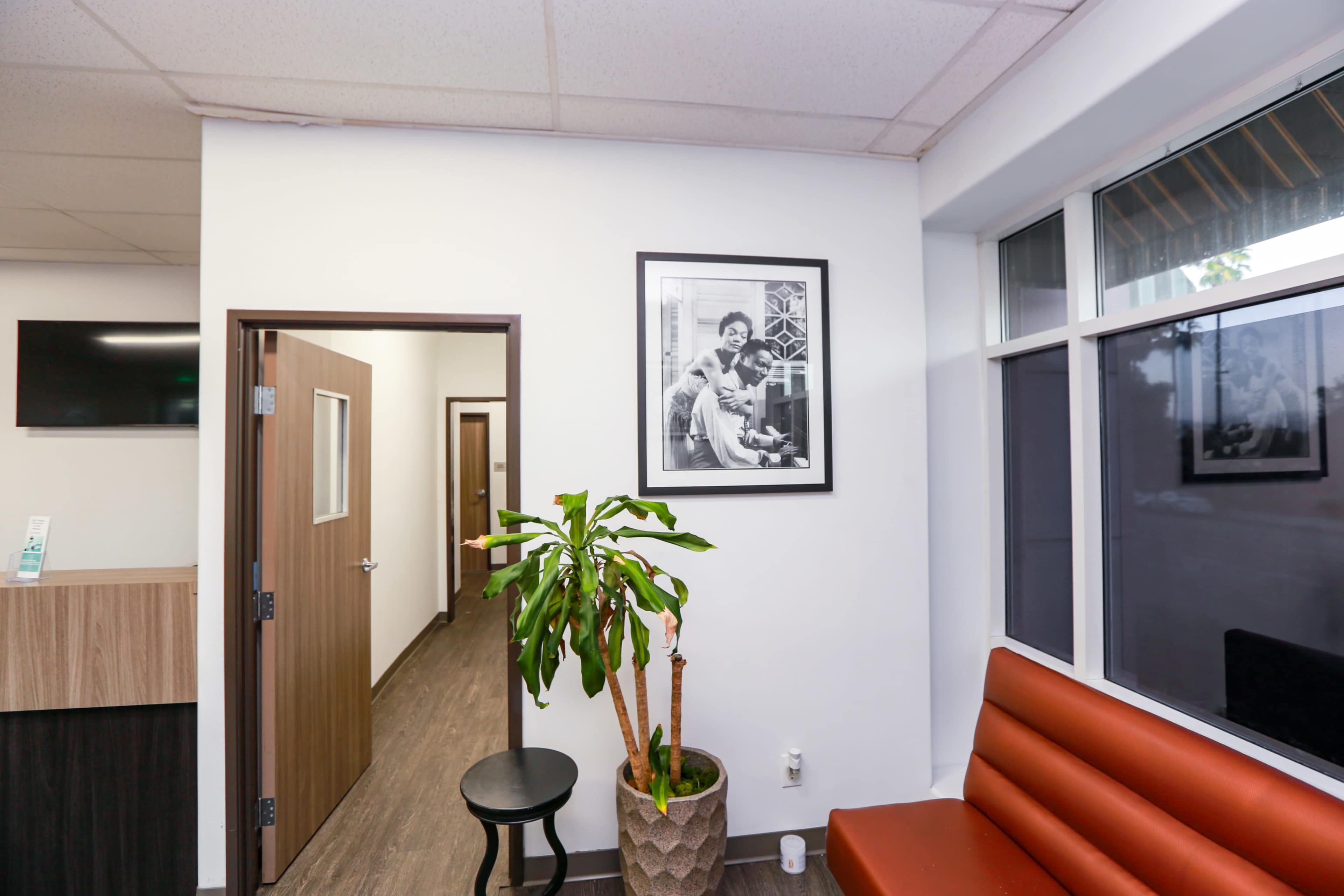 The image shows a waiting area with a brown bench, a small round table, a potted plant, and a black-and-white photograph on the wall.