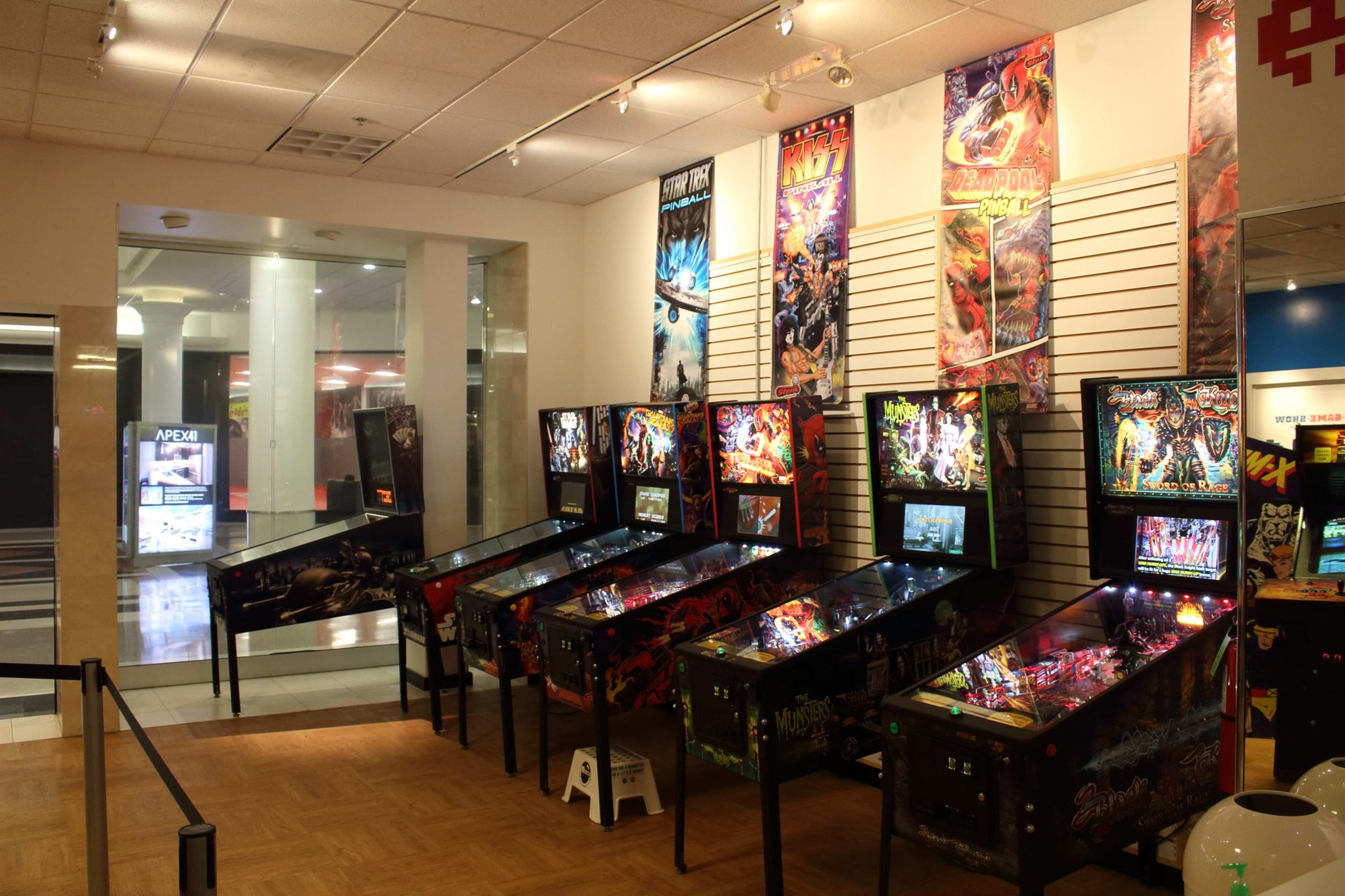 The image shows a row of pinball machines displayed in an arcade, with colorful posters hanging on the walls above them.