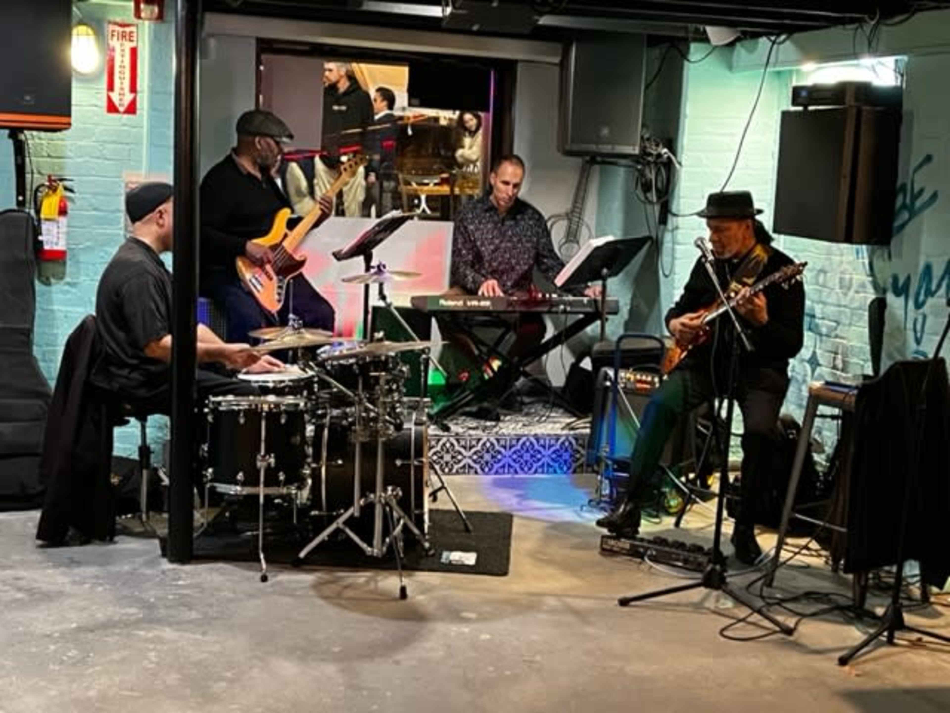 A four-member band performs in a dimly lit basement venue, with a drummer, bassist, keyboardist, and guitarist.