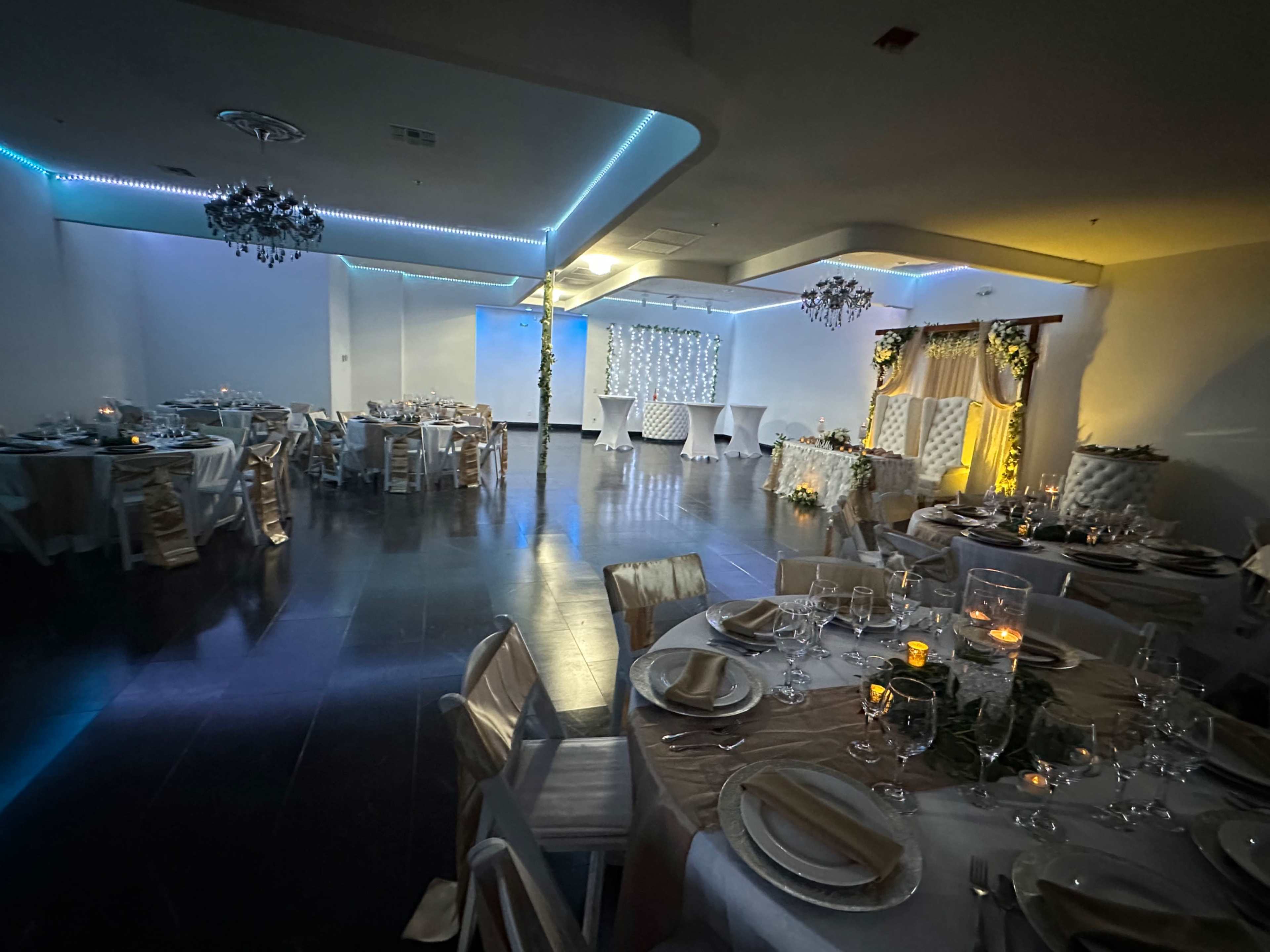 The image shows an elegantly arranged event space with round tables set for a banquet, decorated with white tablecloths and soft lighting, featuring a backdrop with floral accents.