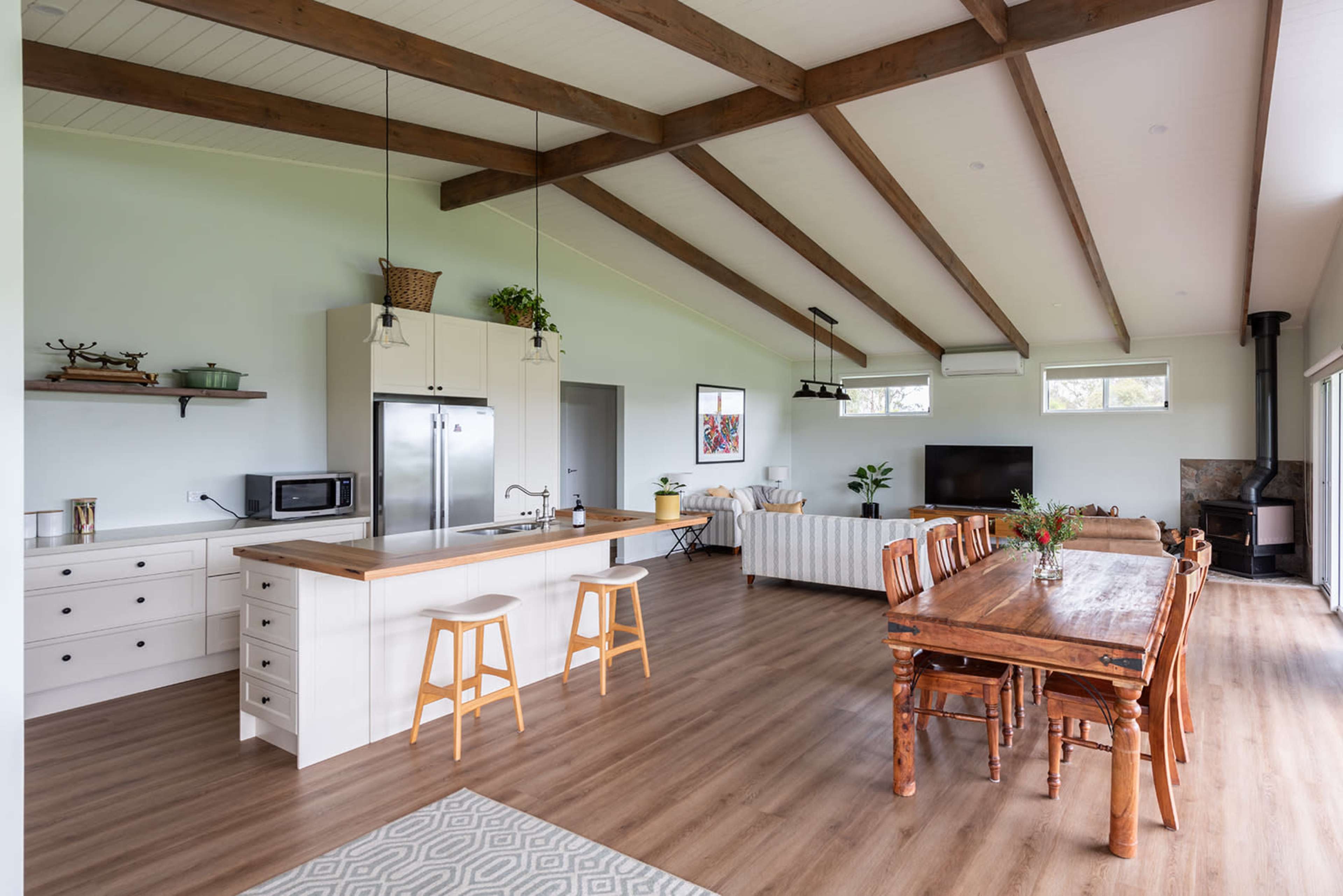 The image shows an open-concept kitchen and dining area with wooden beams, a central island, and a long dining table set against a backdrop of large windows.