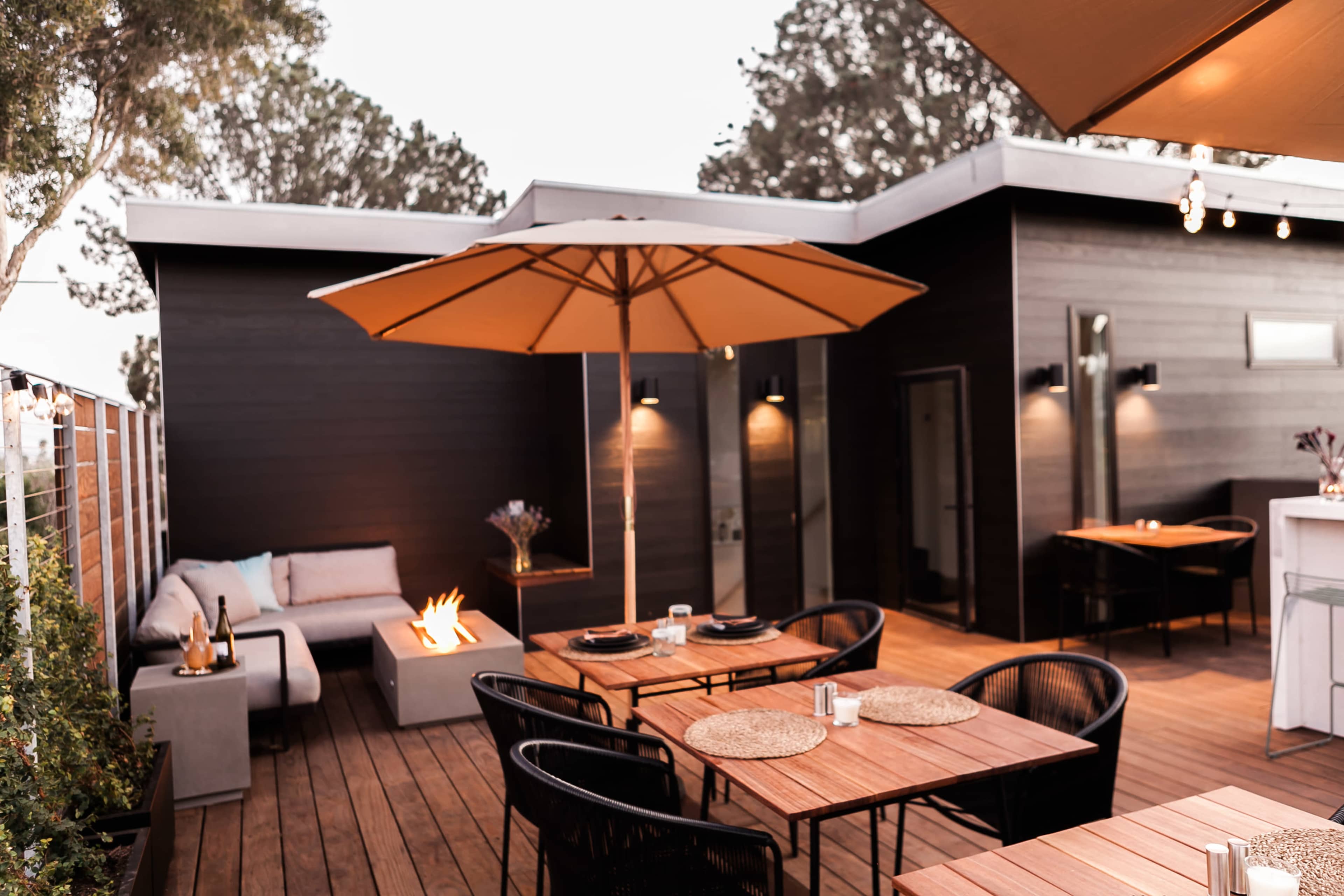 A modern outdoor patio features wooden decking, dining tables, an umbrella, and a cozy seating area with a fire feature.