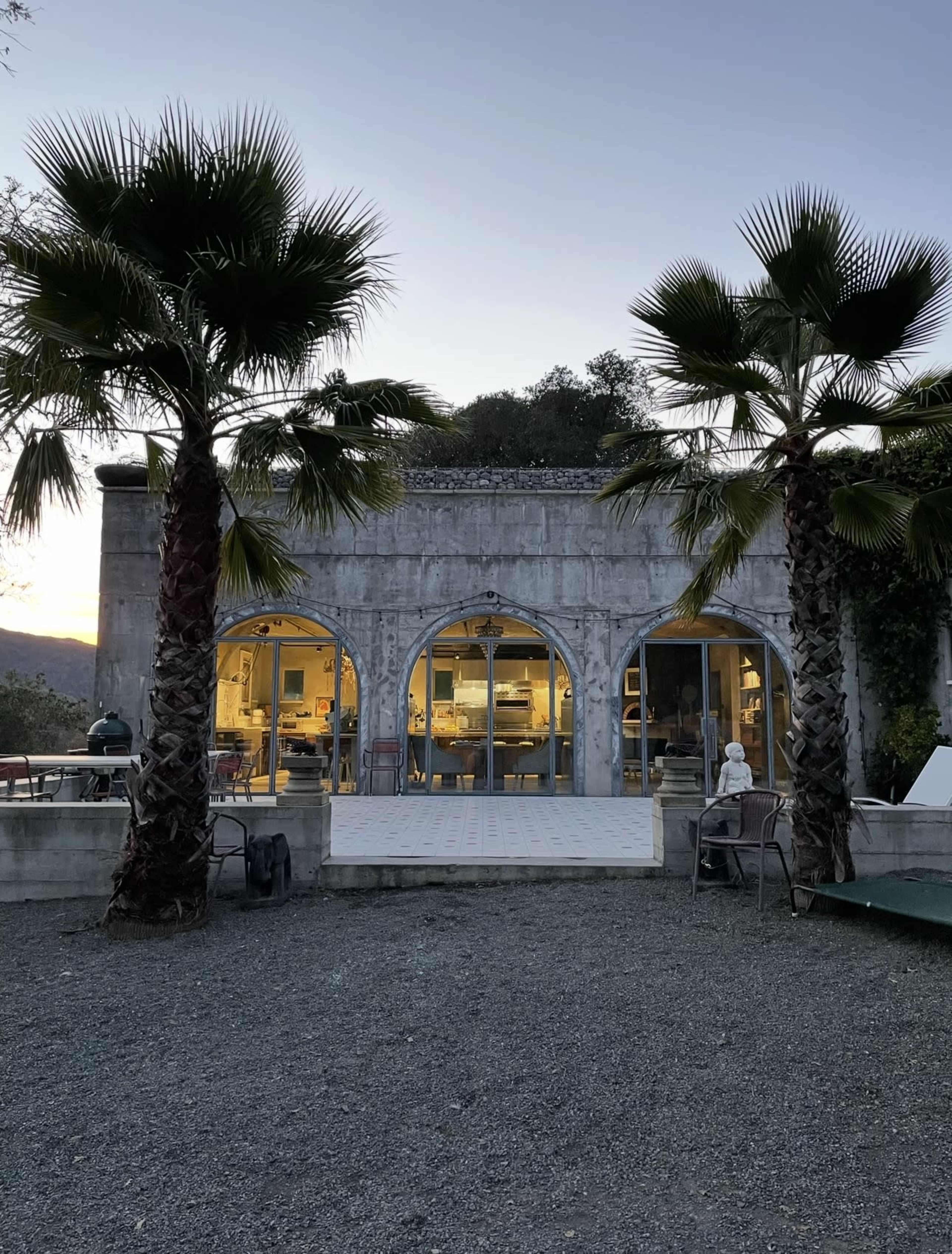 The image shows a modern, curved-arch building with large glass windows, flanked by two palm trees and set in a gravel yard.