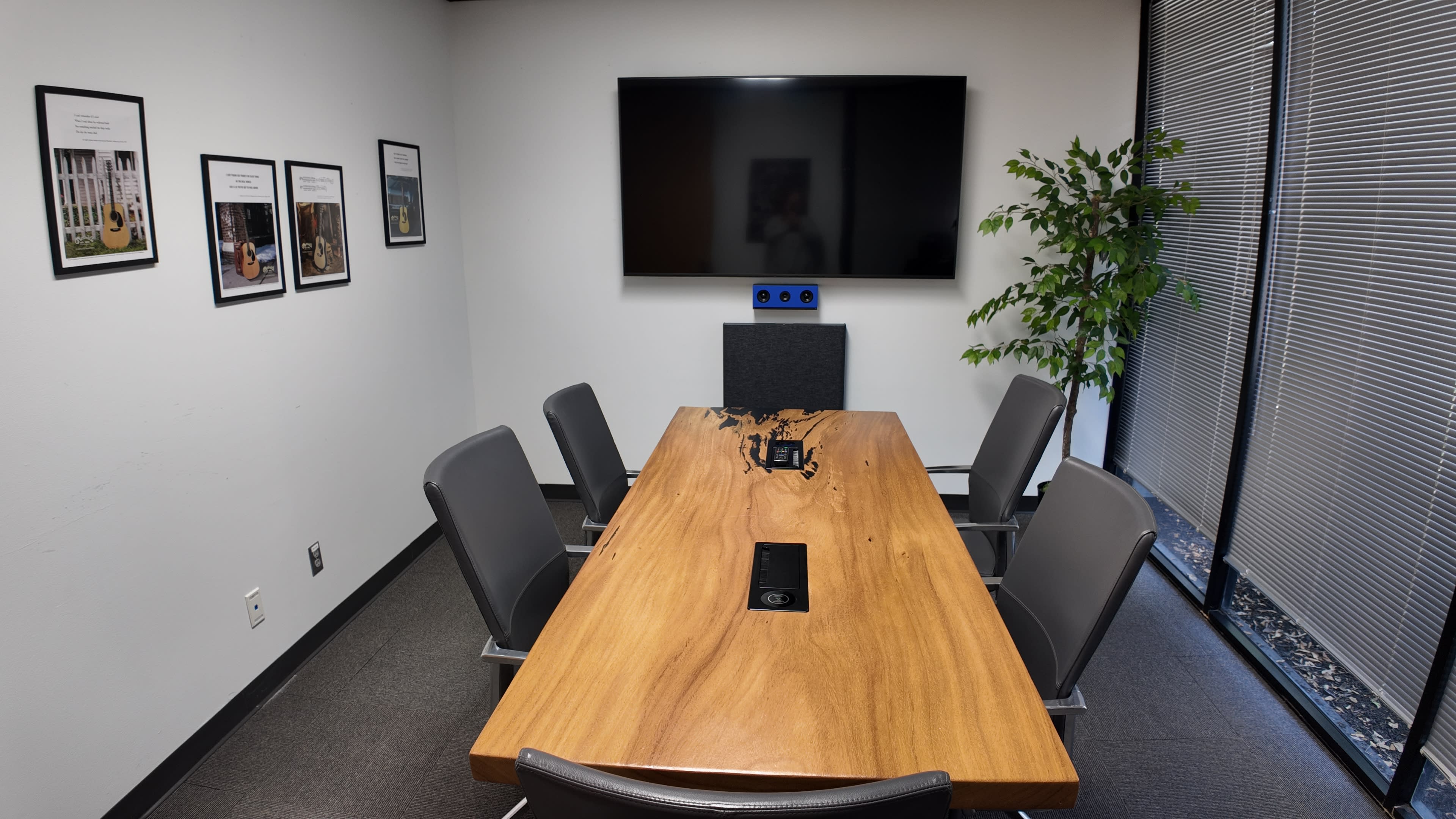 A modern conference room features a large wooden table surrounded by six chairs, with a TV mounted on the wall and framed photos displayed nearby.