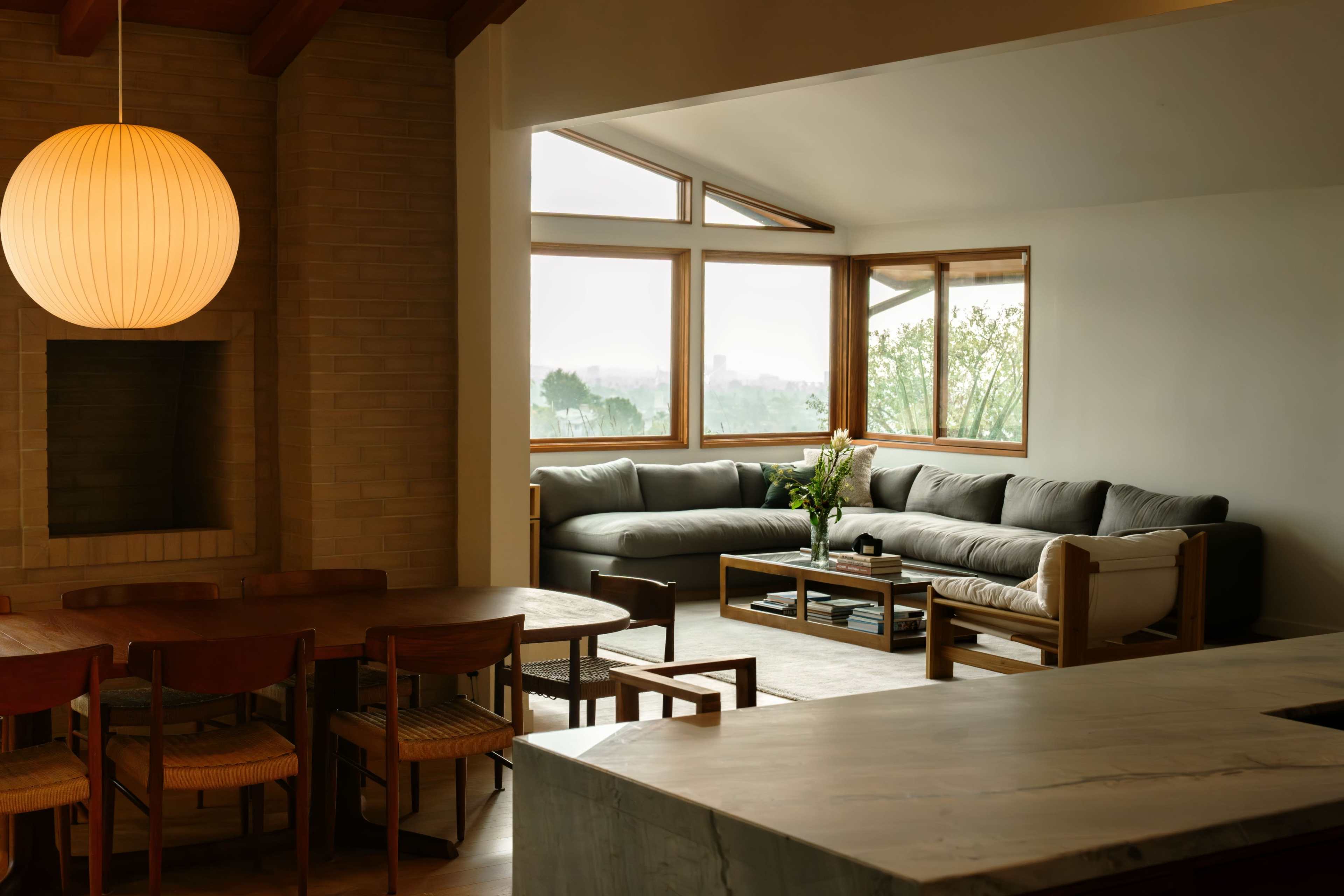 A modern living room features a large gray sectional sofa, a wooden dining table, and a view through large windows.