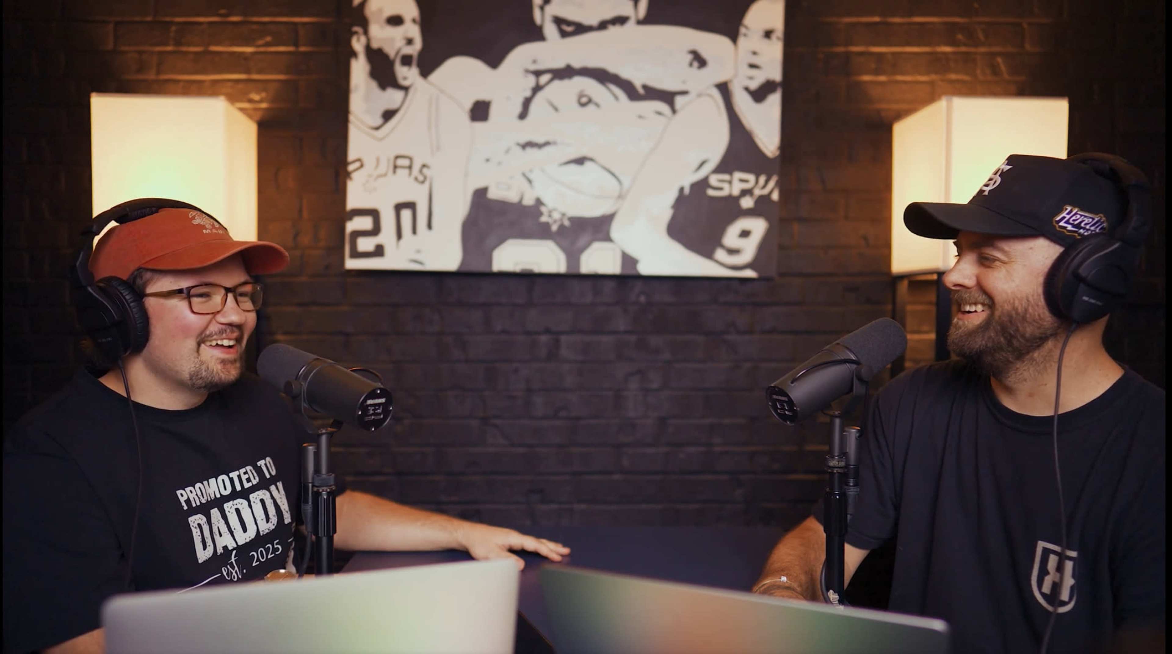 Two men are sitting at a table with microphones, engaged in conversation, while a black-and-white sports-themed mural hangs on the wall behind them.