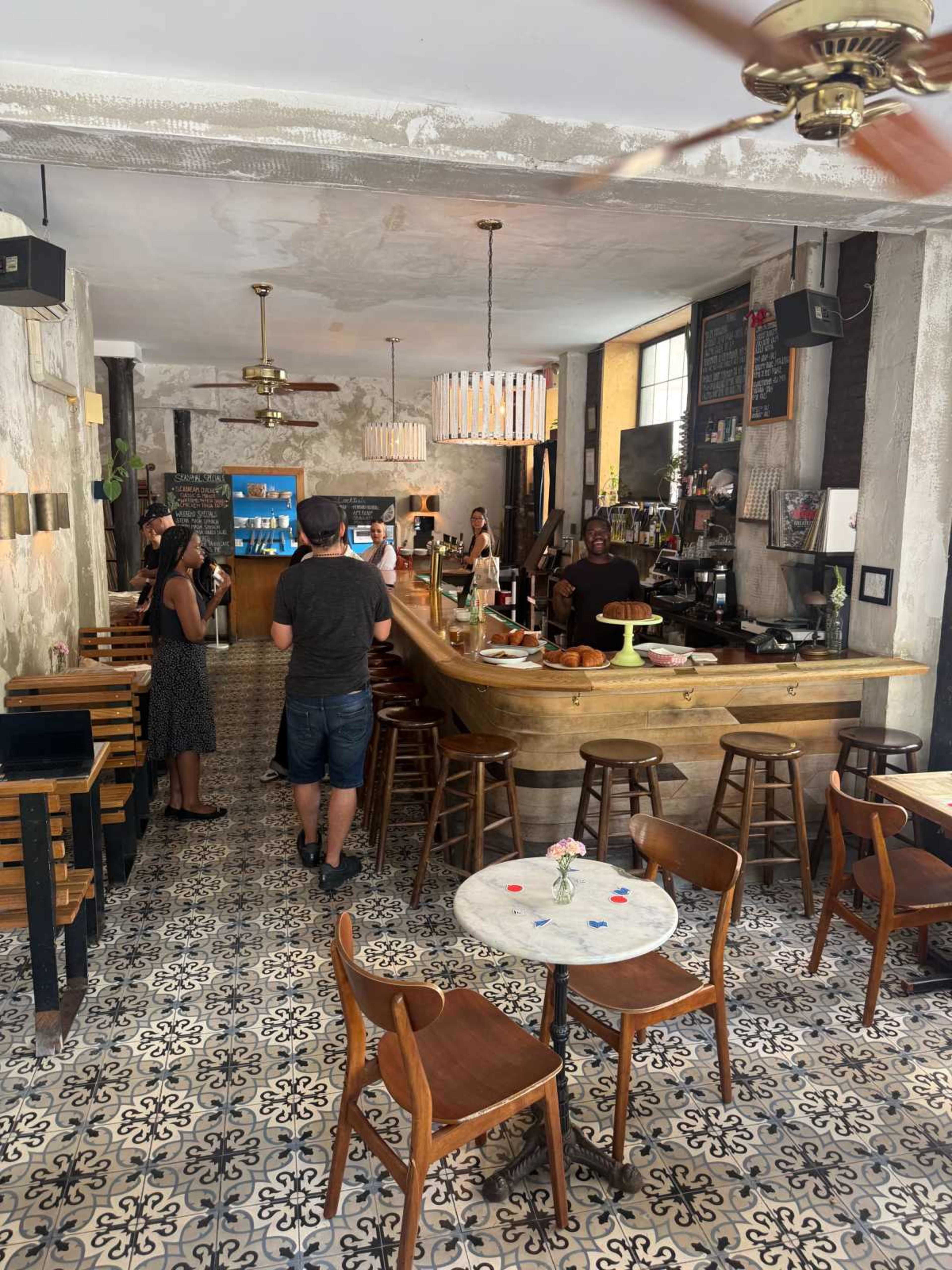 The image shows a cozy cafe interior with a wooden bar, tiled flooring, and several patrons seated at tables.