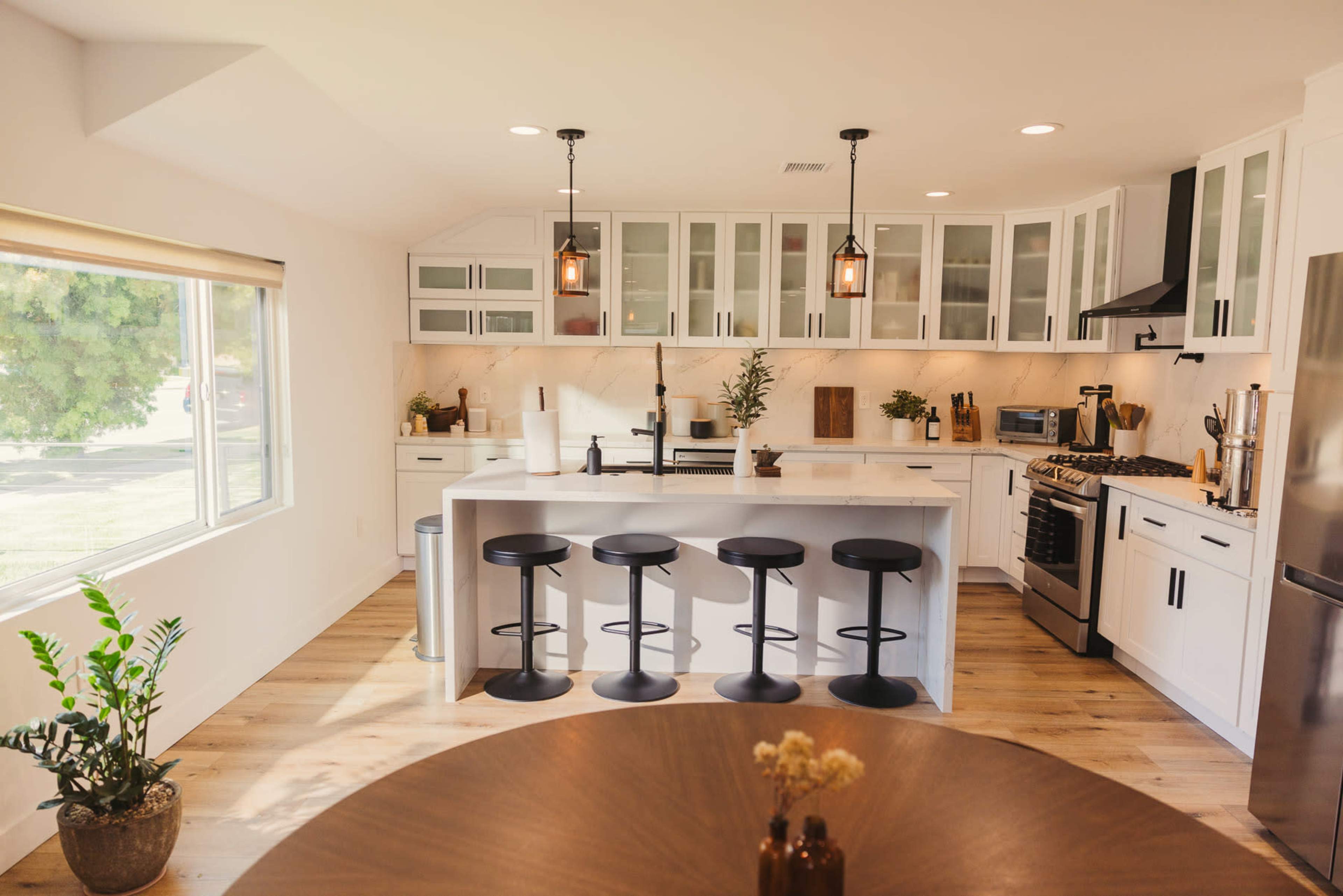 The image shows a modern kitchen with white cabinetry, a central island with black bar stools, and large windows allowing natural light.