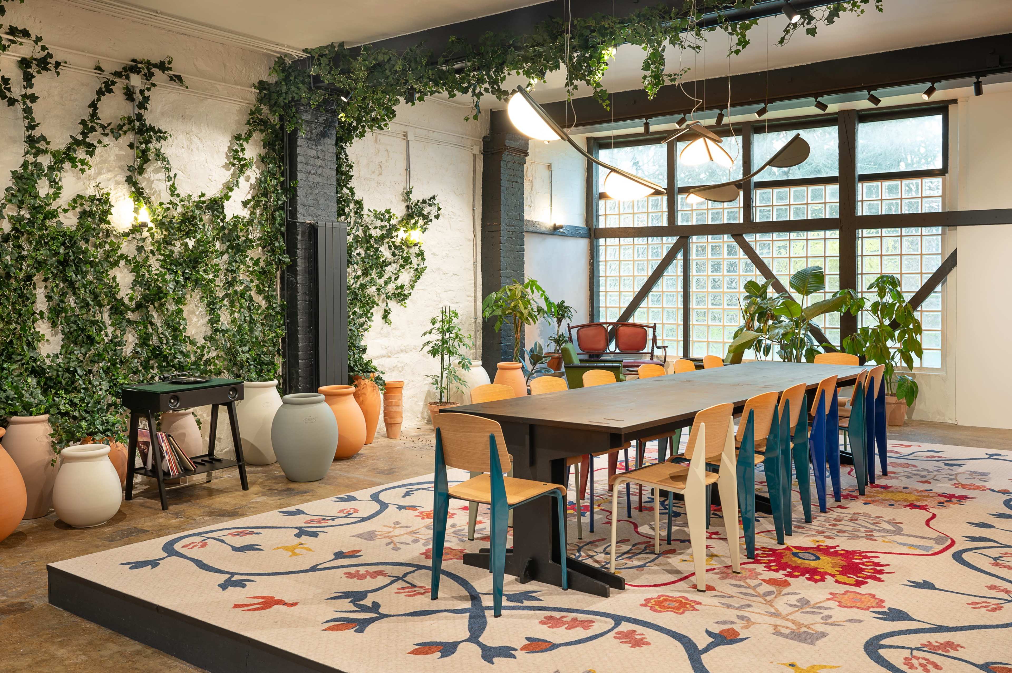 The image shows a spacious dining area featuring a long table surrounded by colorful chairs, with large windows adorned by plants and a decorated rug on the floor.