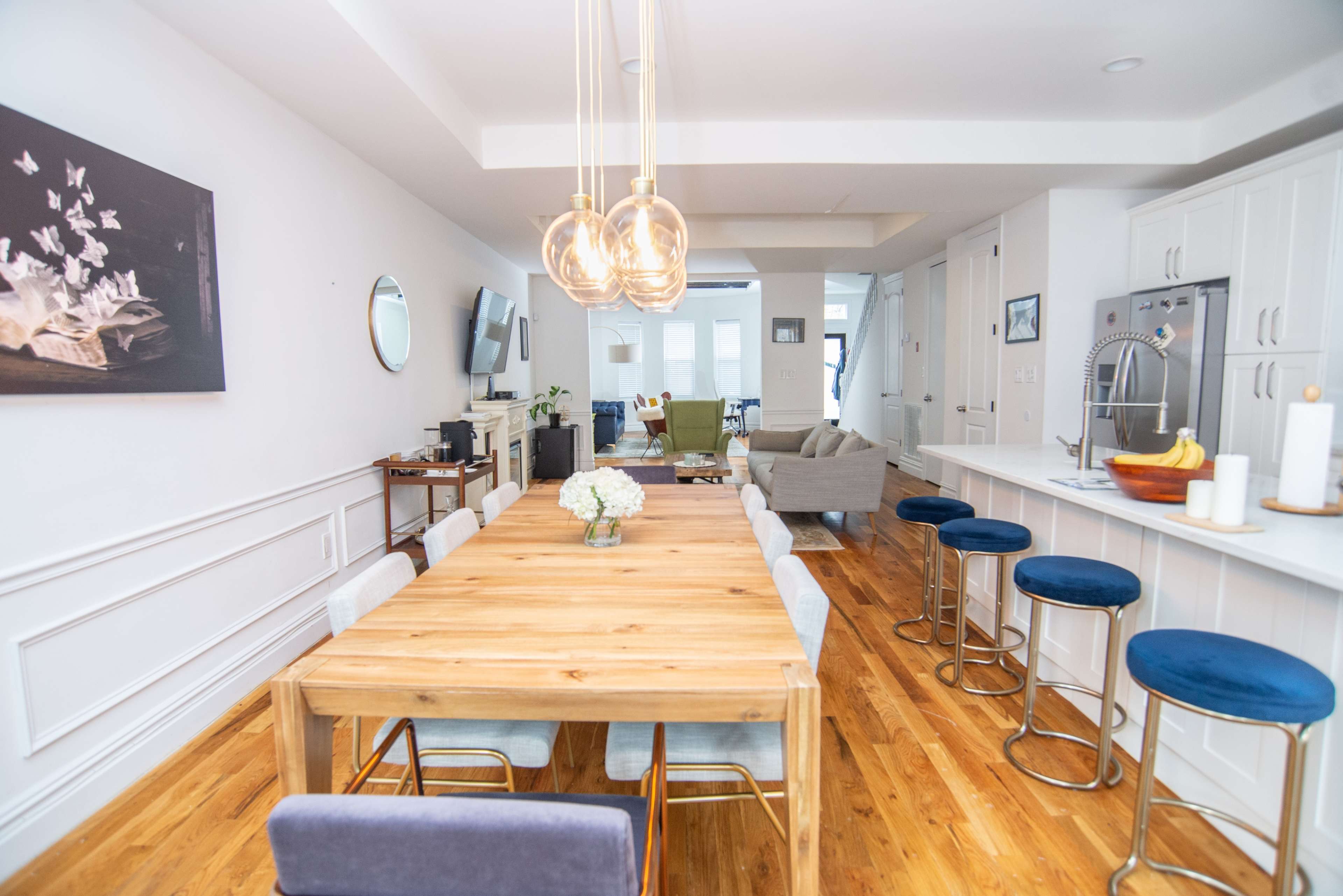 A modern dining area with a large wooden table surrounded by upholstered chairs, leading into a bright living space featuring a sofa and television.