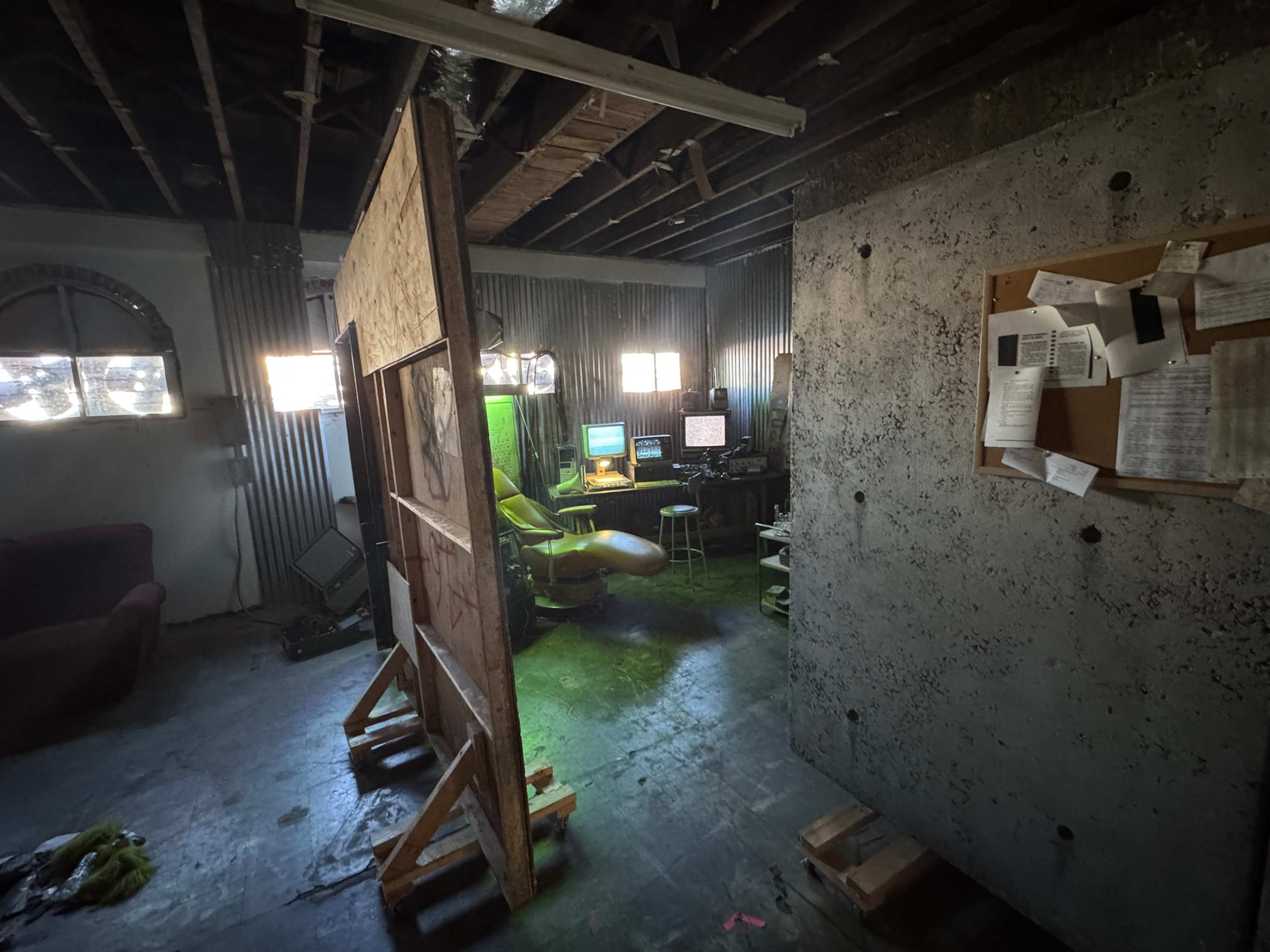 The image shows a dimly lit room with a green office chair facing multiple computer screens, separated by a wooden partition and surrounded by bare walls and scattered paperwork.
