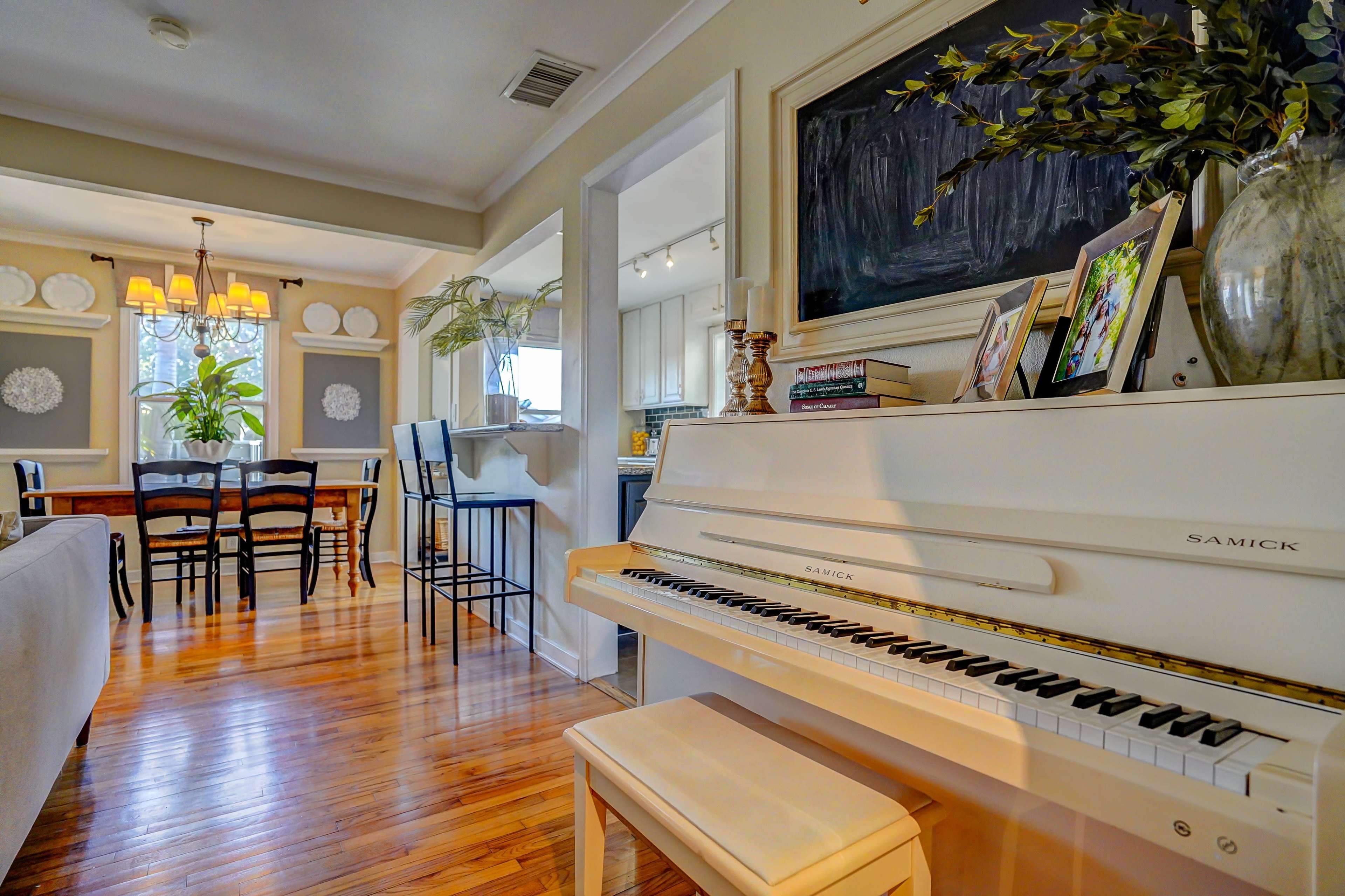 A white piano sits in the foreground of a bright living space that includes a dining area and an adjacent kitchen.