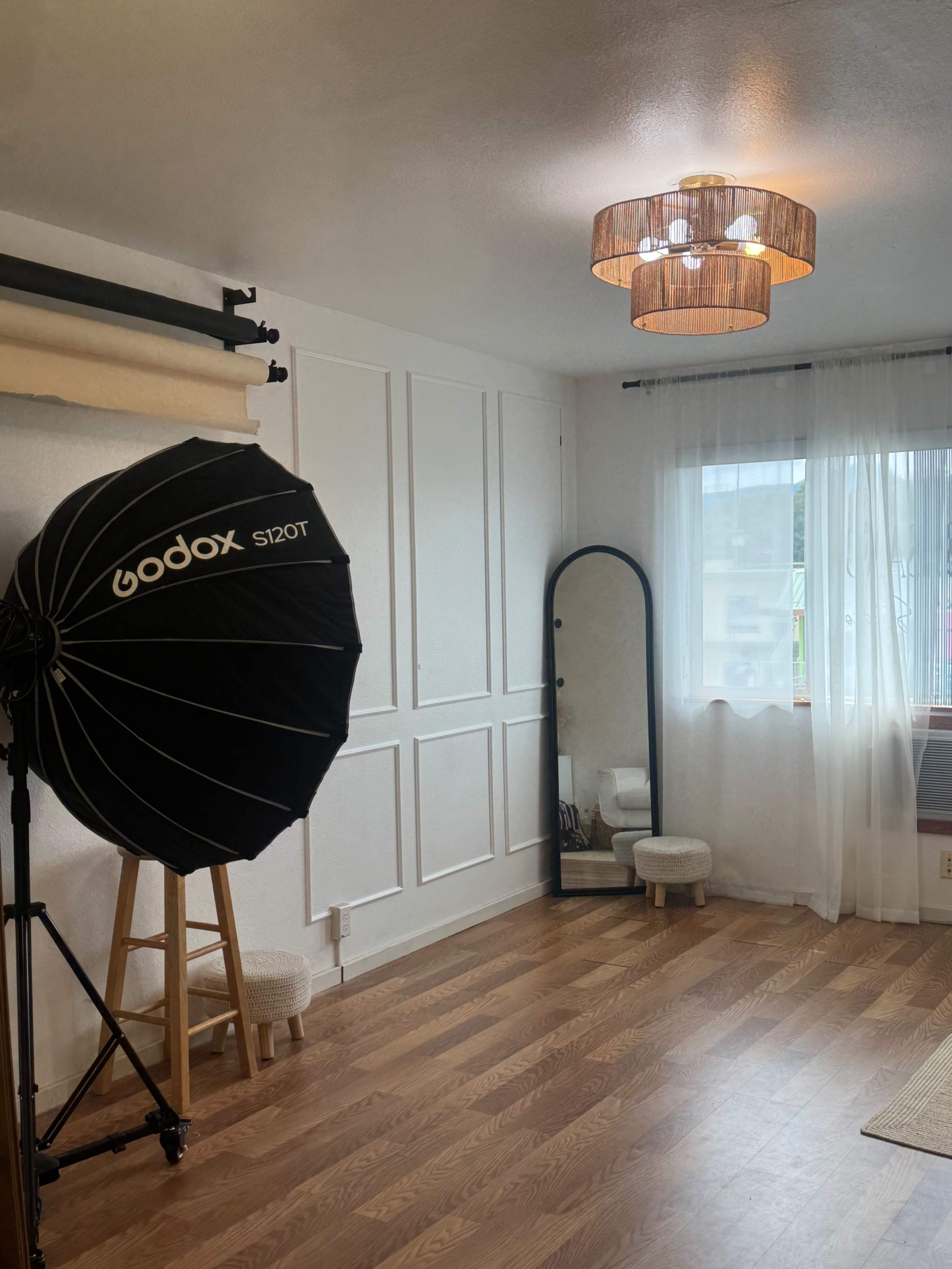The image shows a well-lit room with wooden flooring, featuring a large black photography reflector, a mirror, and a chandelier.