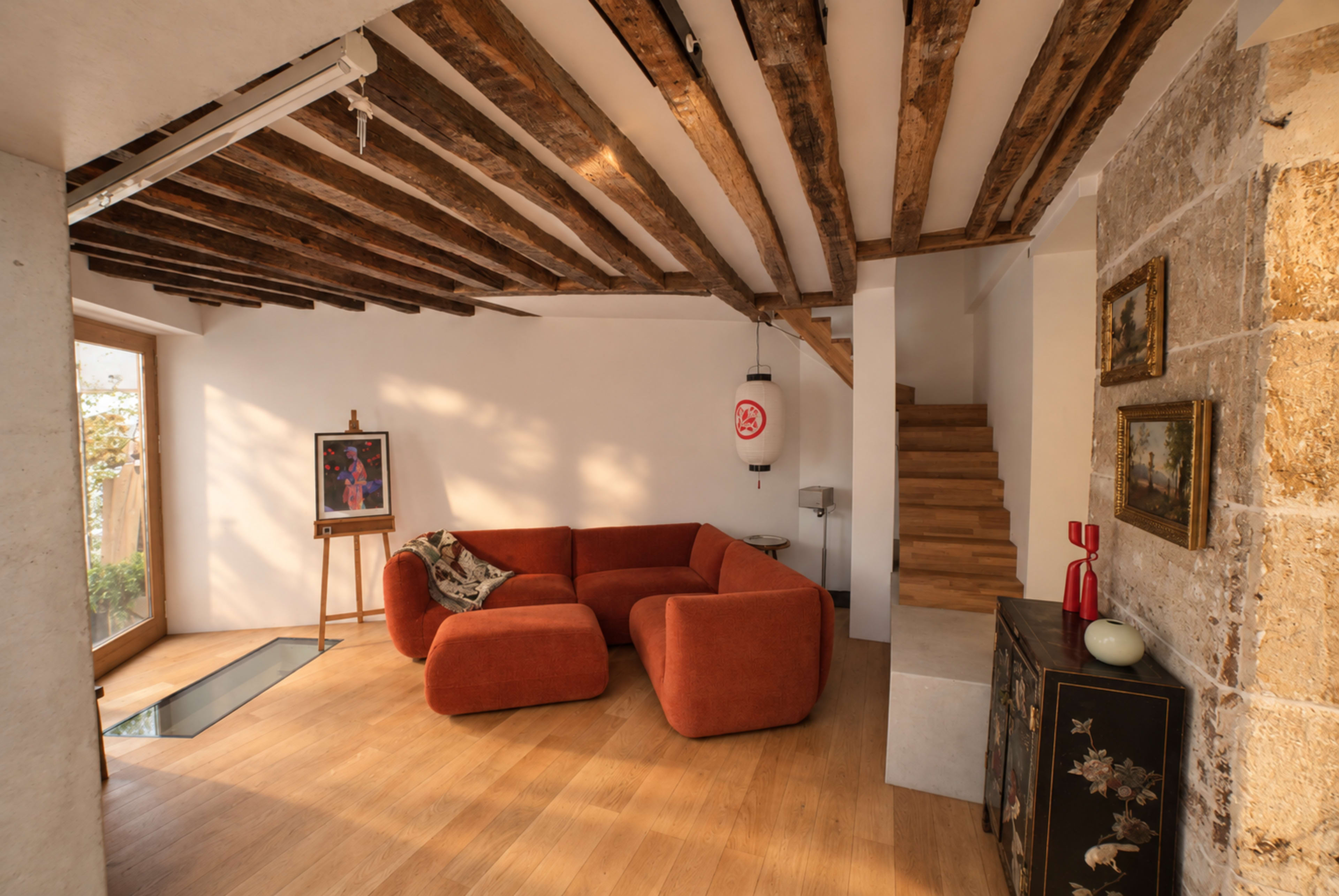 The image shows a modern living room with a large orange sectional sofa, wooden beams on the ceiling, a staircase in the background, and a glass panel in the floor.