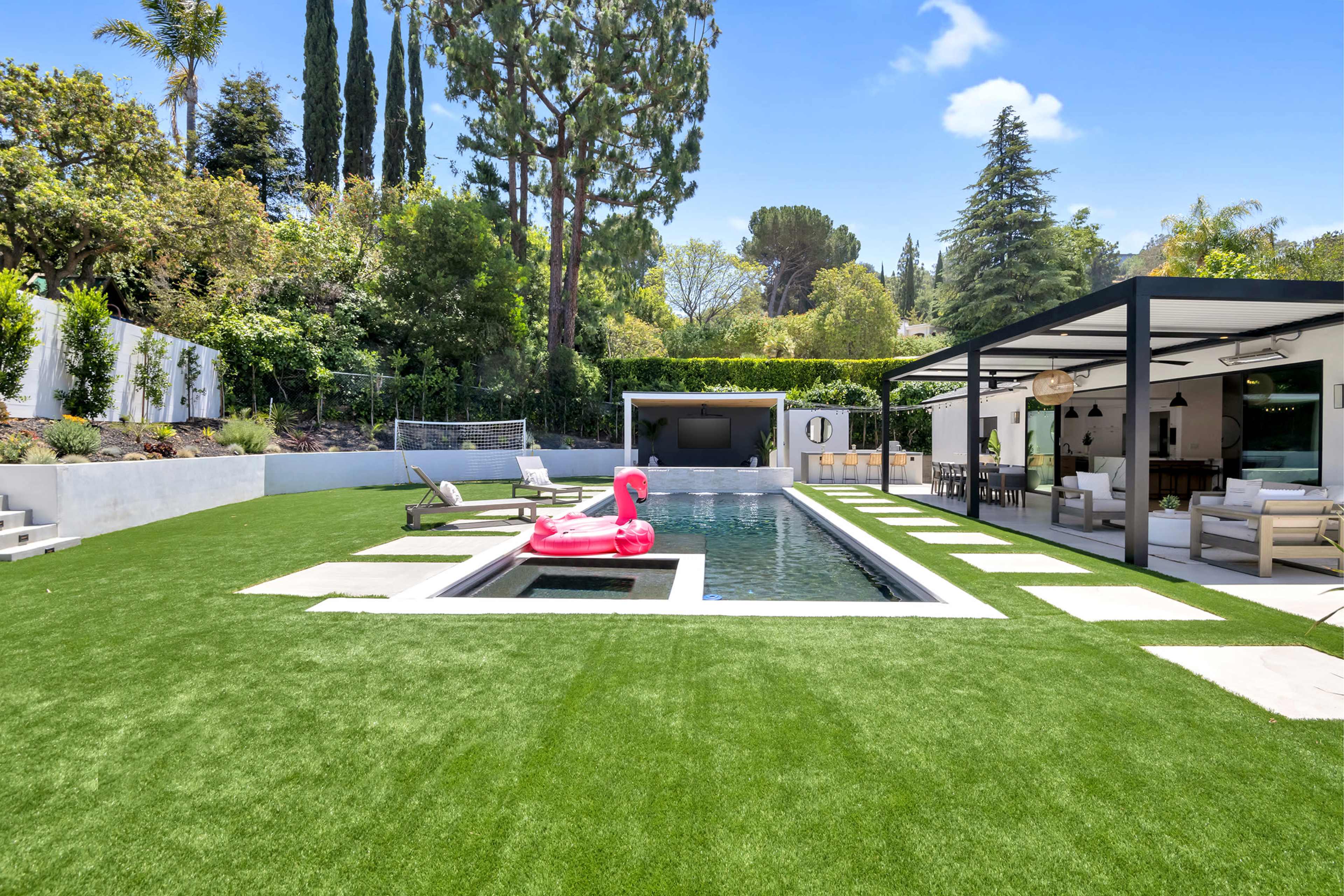 A backyard featuring a swimming pool surrounded by artificial grass, lounge chairs, and a covered seating area with a television.