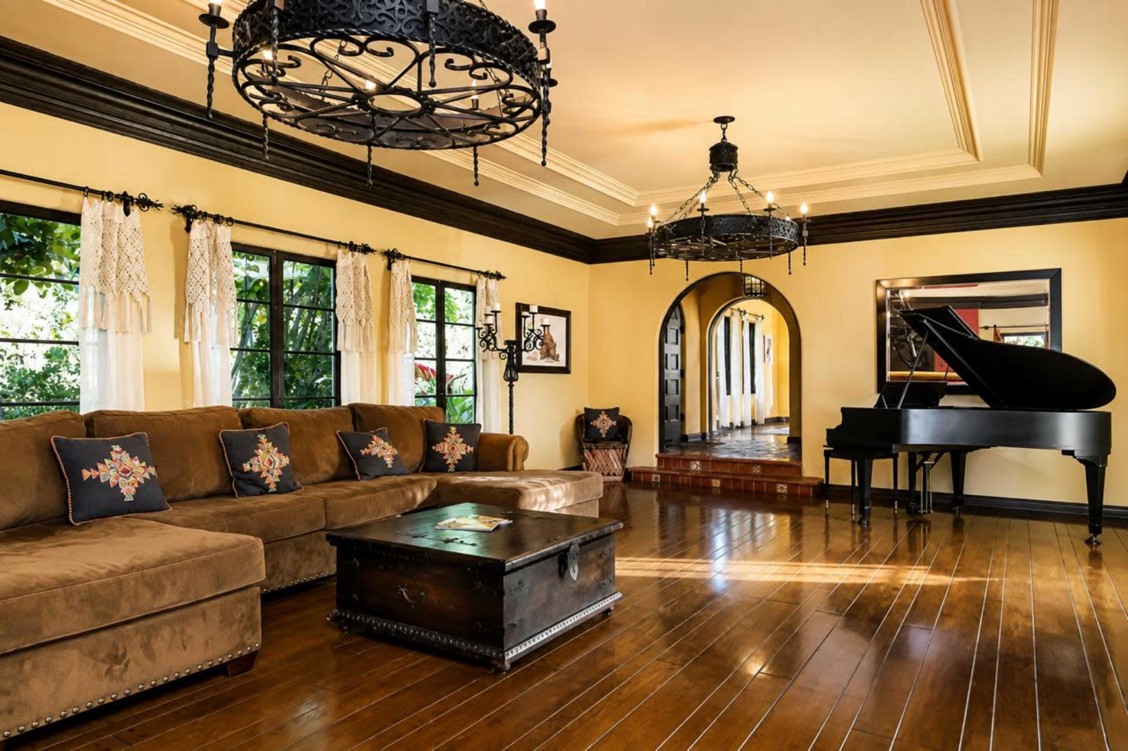 The image shows a spacious living room featuring a large brown sectional sofa, a black grand piano, and two ornate chandeliers, with windows that reveal greenery outside.