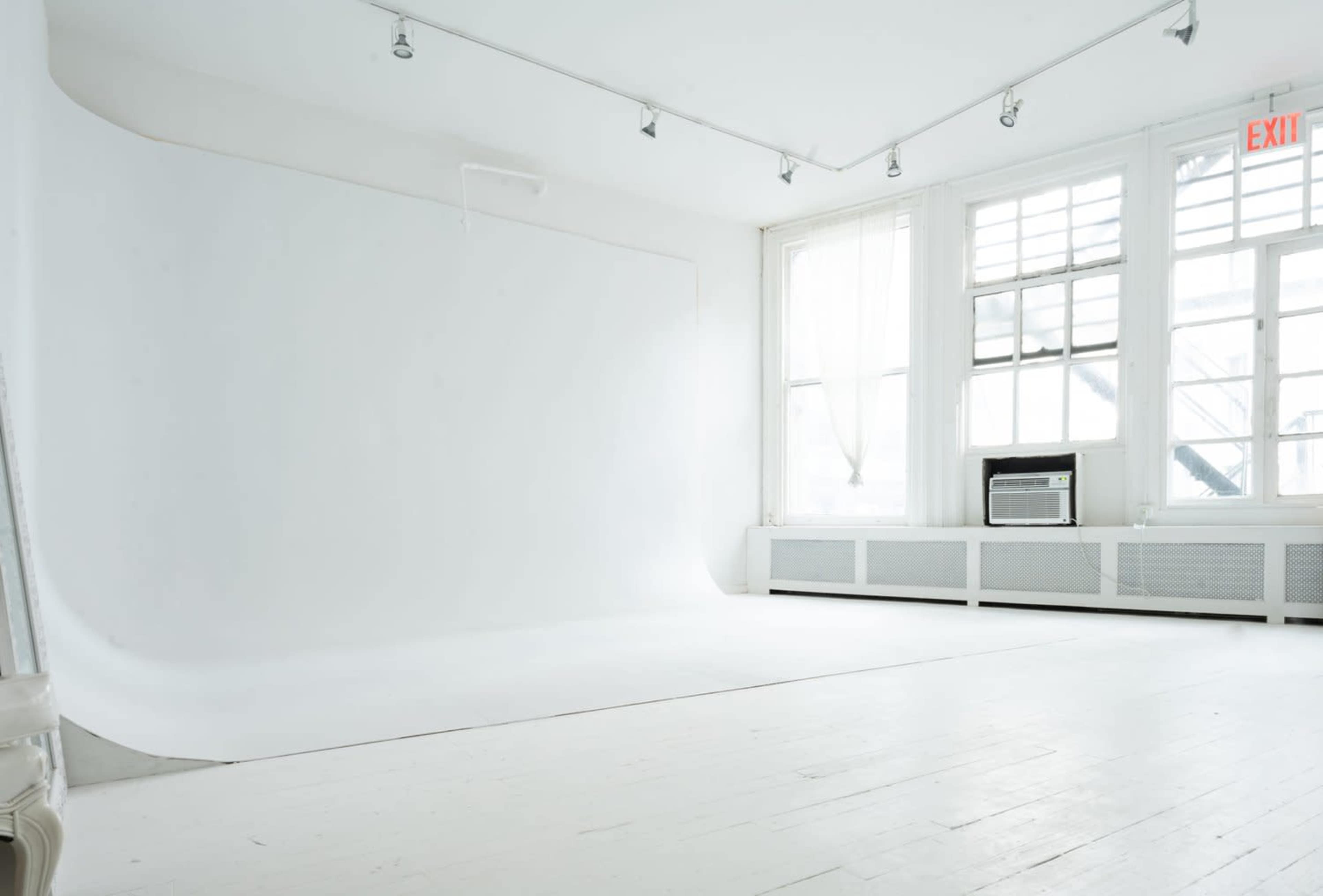 A minimalist photography studio with a seamless white backdrop and large windows allowing natural light to enter.