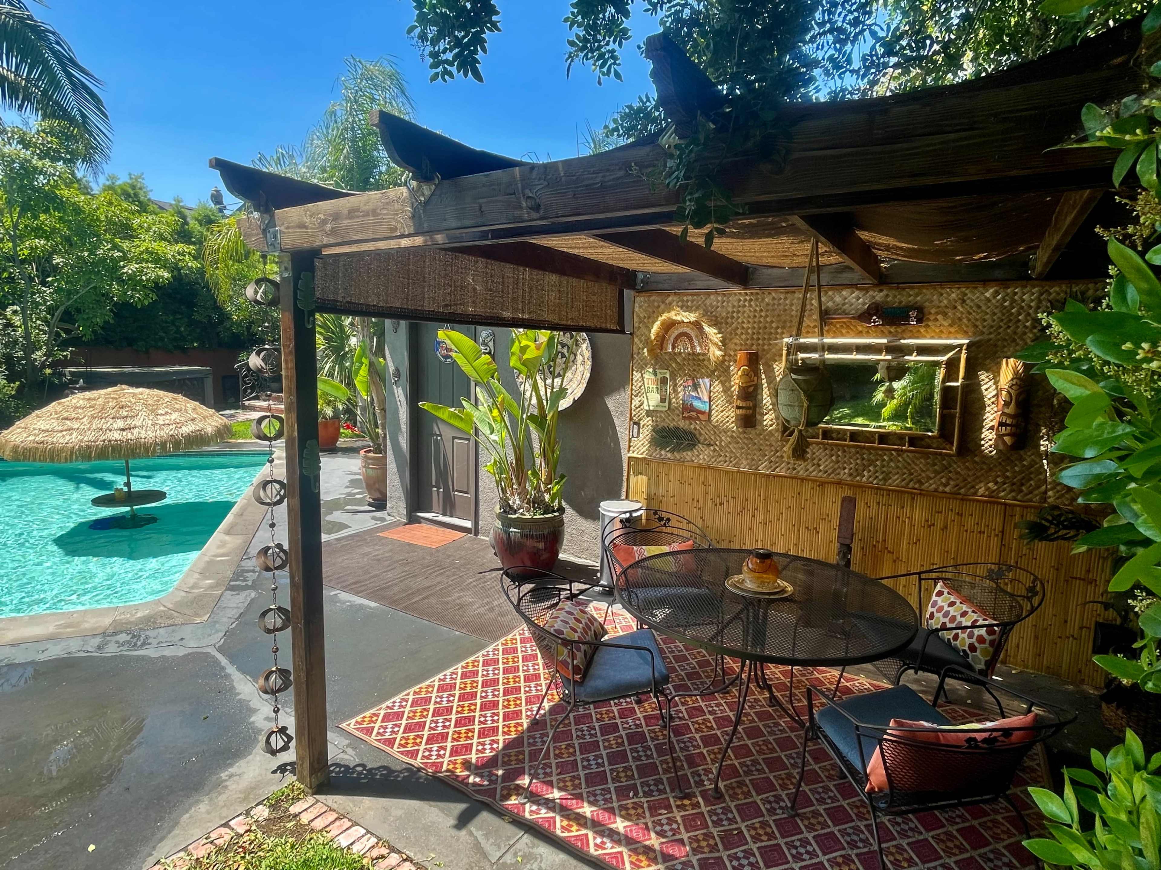 A shaded outdoor seating area with a table and chairs overlooks a swimming pool surrounded by greenery.