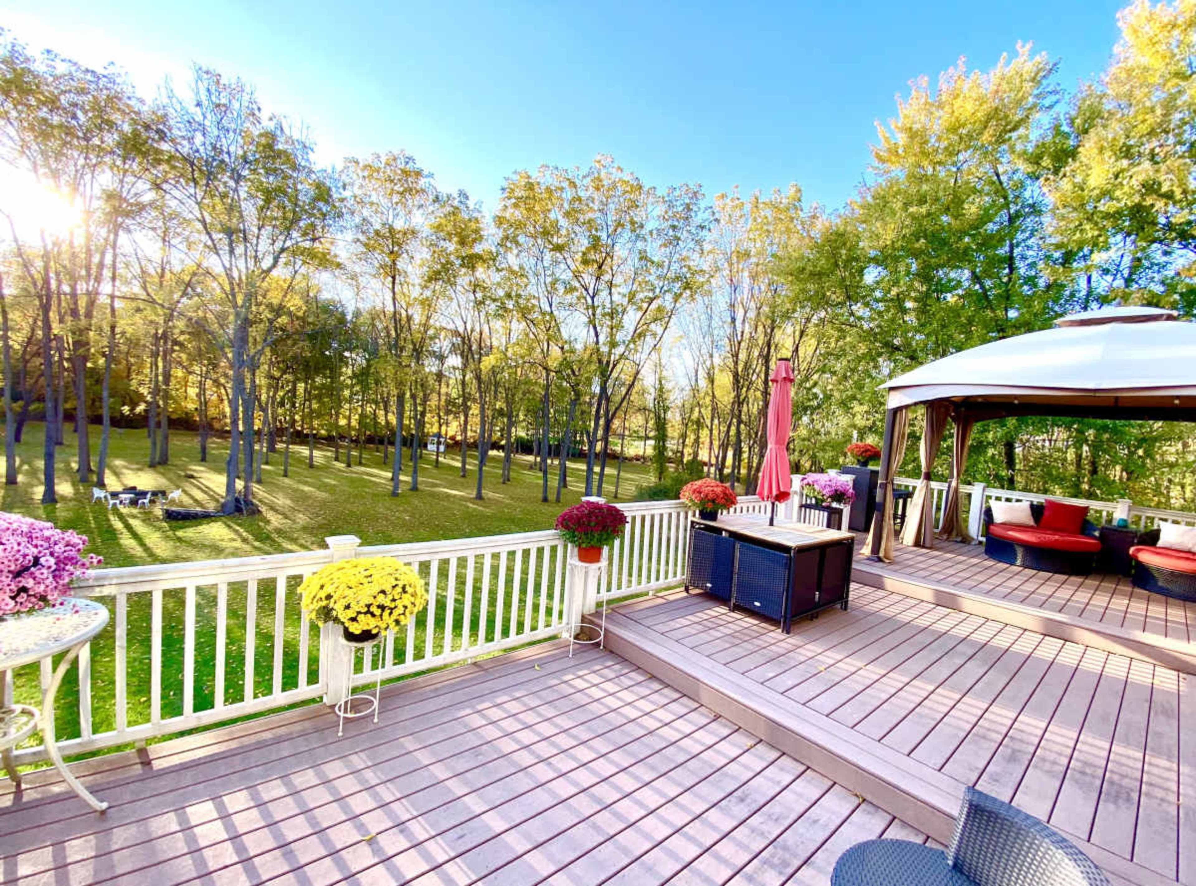 A spacious wooden deck overlooks a lush green yard surrounded by trees, with colorful flower pots and a gazebo on one side.