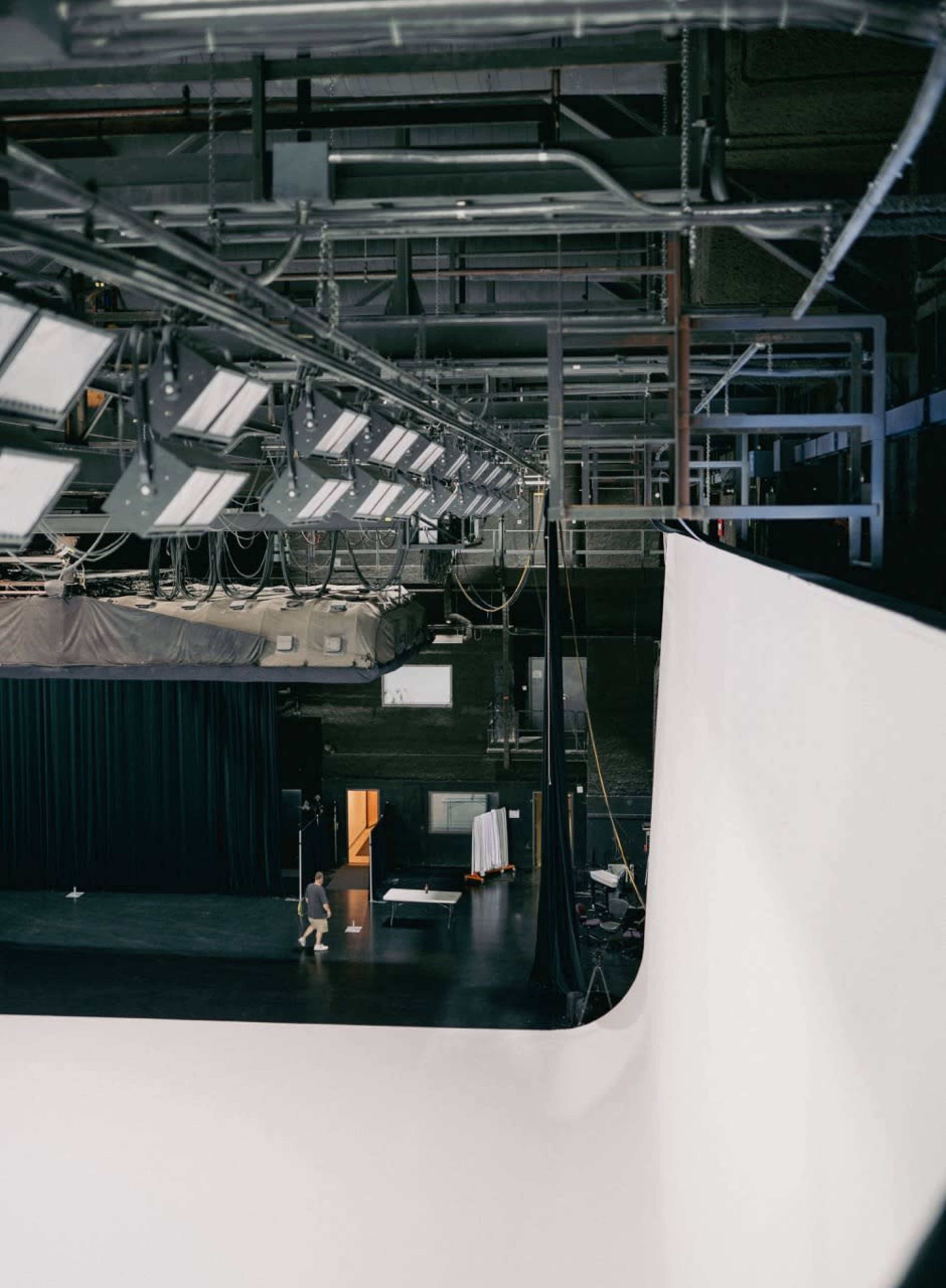 A spacious film studio features a white backdrop, overhead lighting, and various equipment, with a person walking across the floor.
