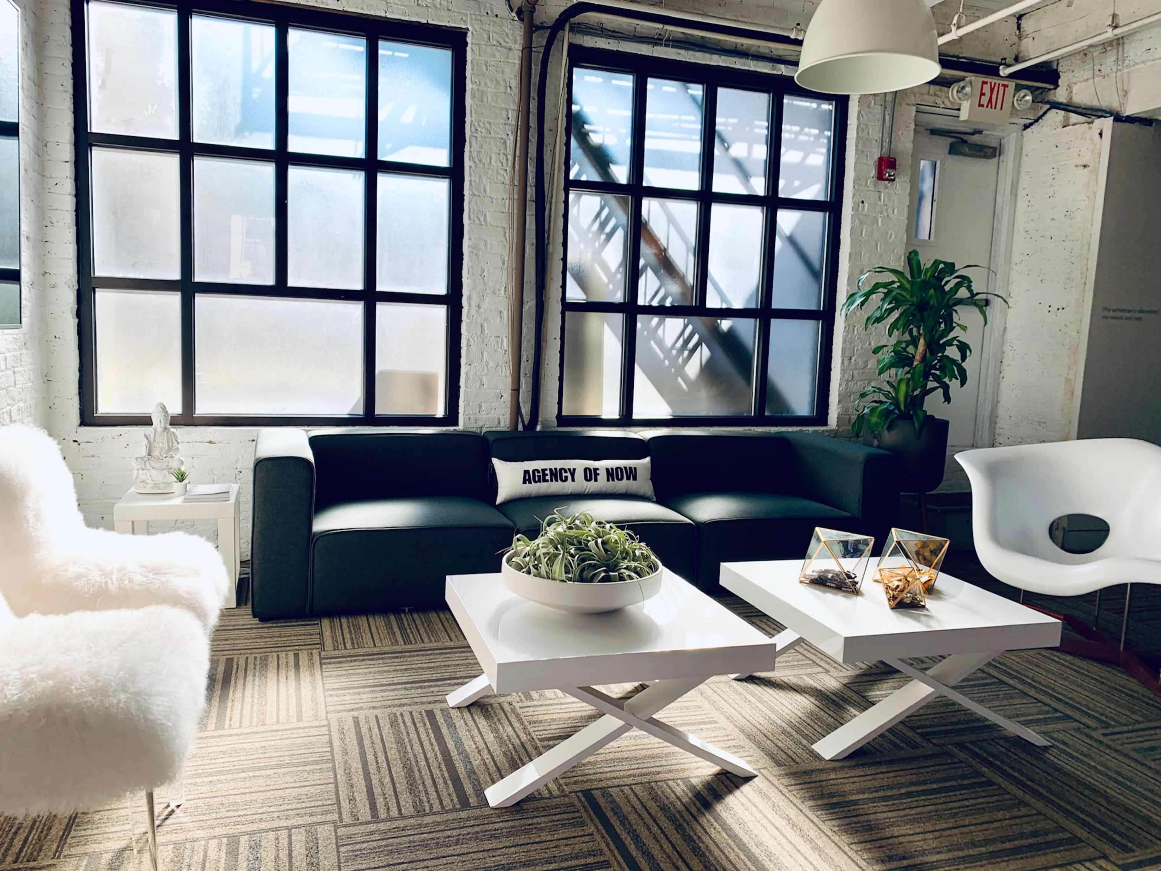 The image shows a modern office lounge area featuring a dark green sofa, white coffee tables, and large windows, with plants and decorative items.