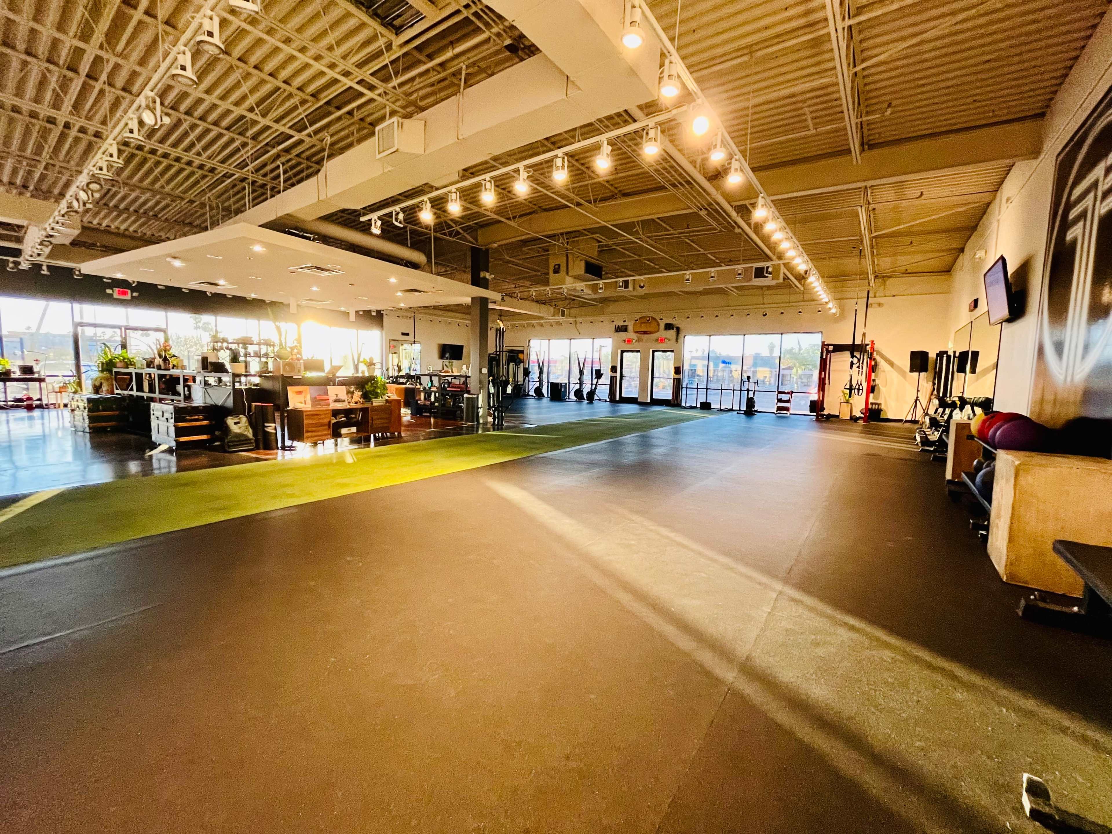 A spacious gym interior with workout areas, natural light streaming in through large windows, and various fitness equipment positioned along the walls.
