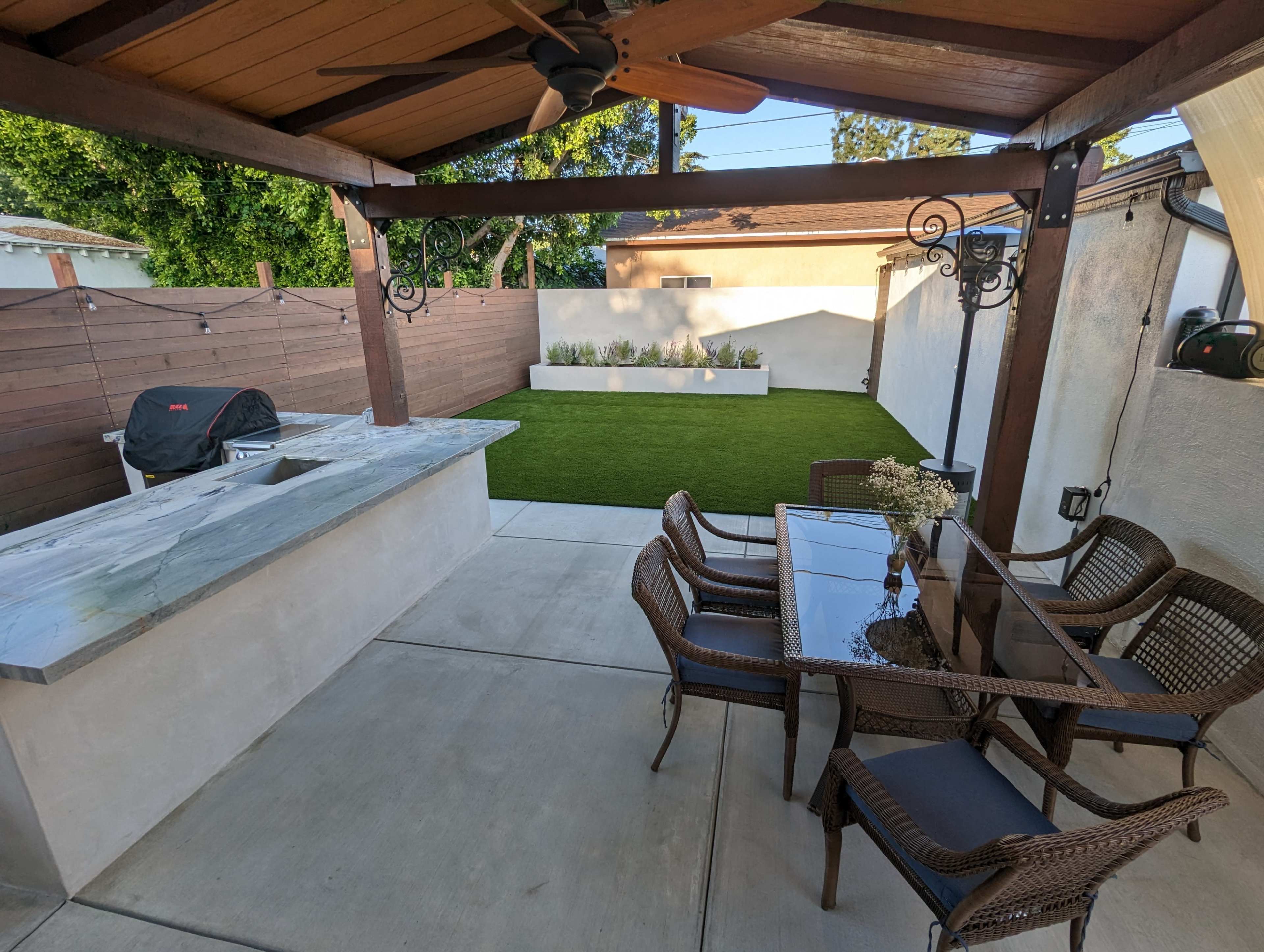 A covered outdoor patio area with a marble countertop, a grill, and a dining set overlooking a small patch of artificial grass.