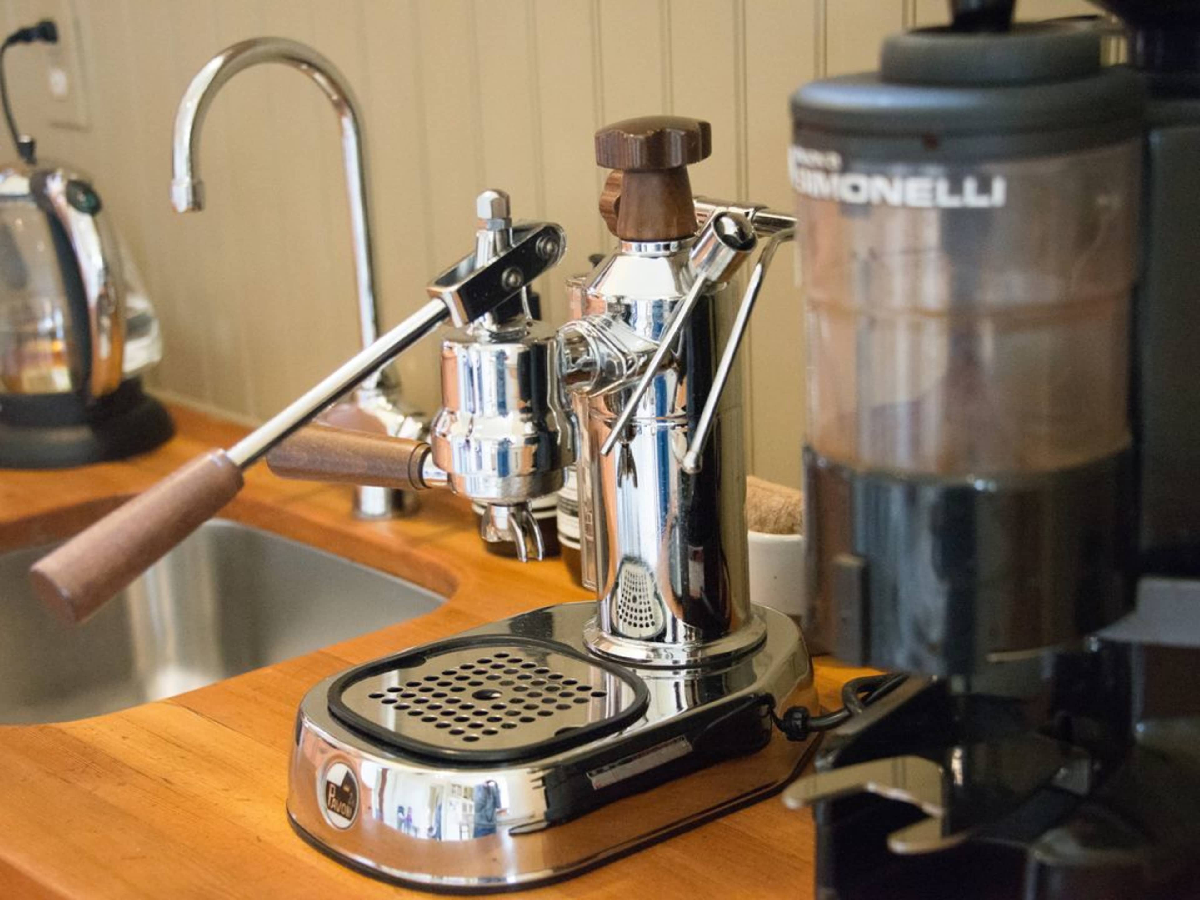 A shiny espresso machine with a wooden handle, positioned on a wooden countertop beside a coffee grinder.