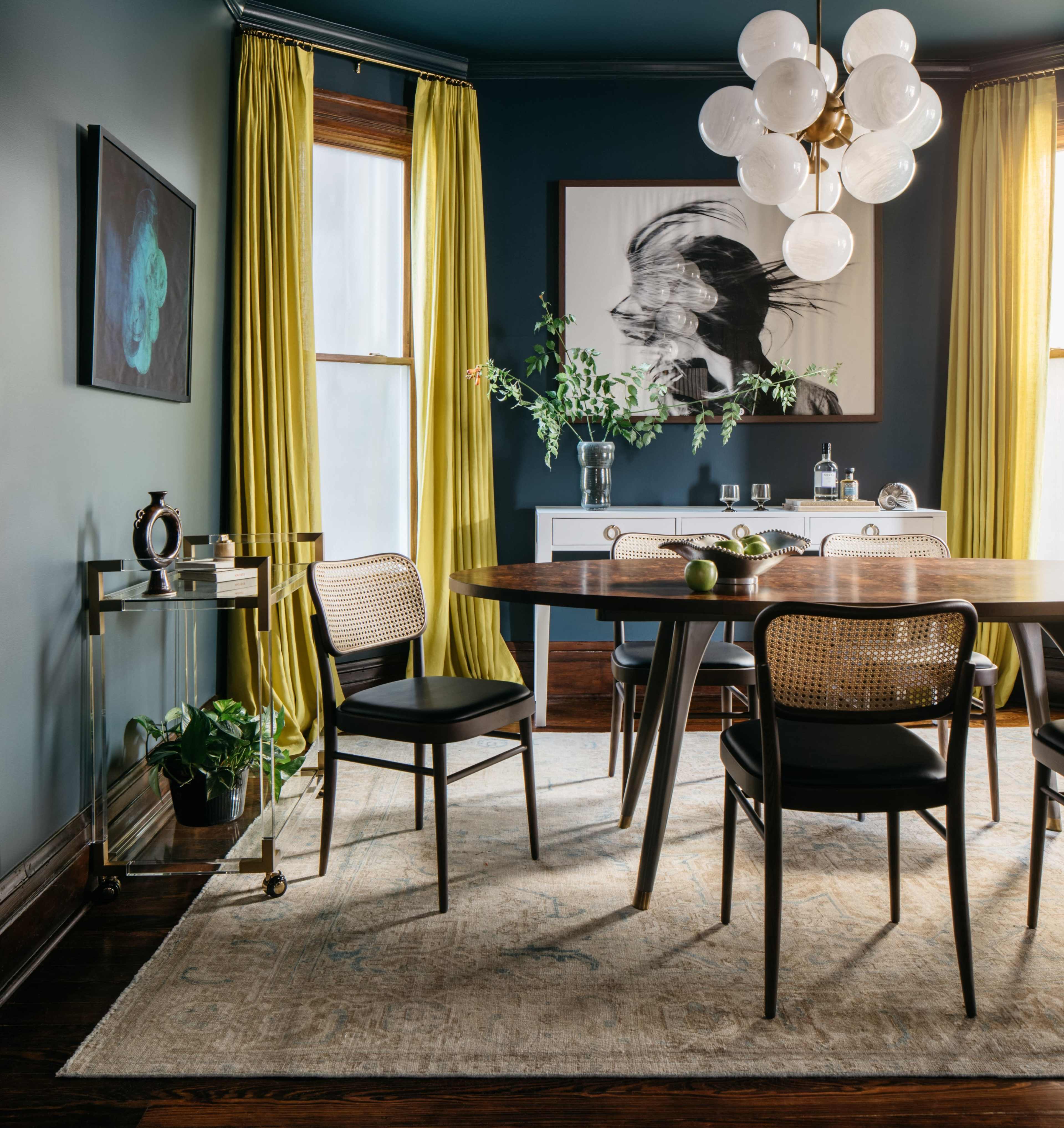 A modern dining room features a round table surrounded by black chairs, a sideboard, and large windows draped with yellow curtains.