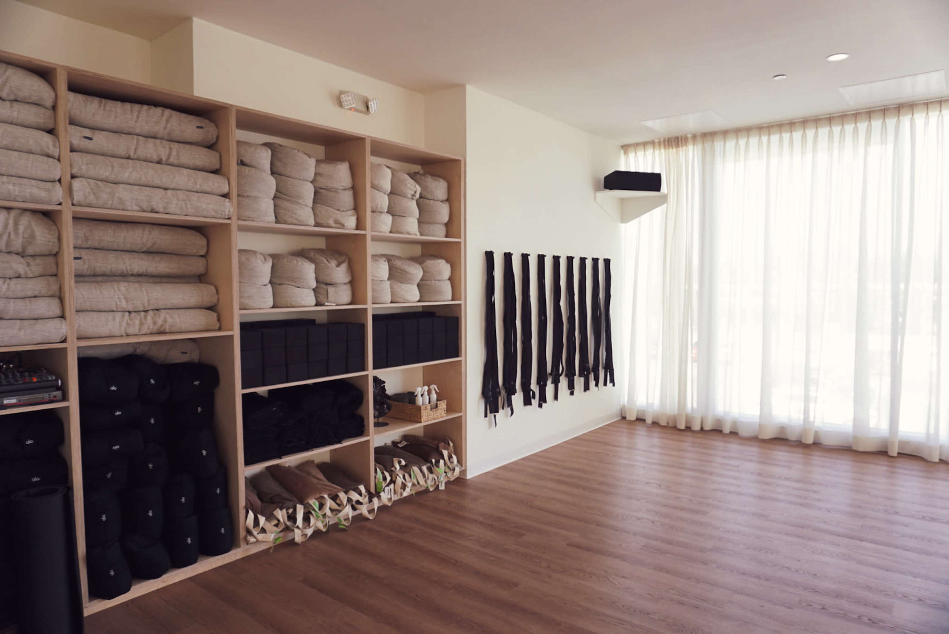 The image shows a minimalist yoga studio with shelves stacked with mat blankets and bolsters, alongside a wall displaying black yoga straps.