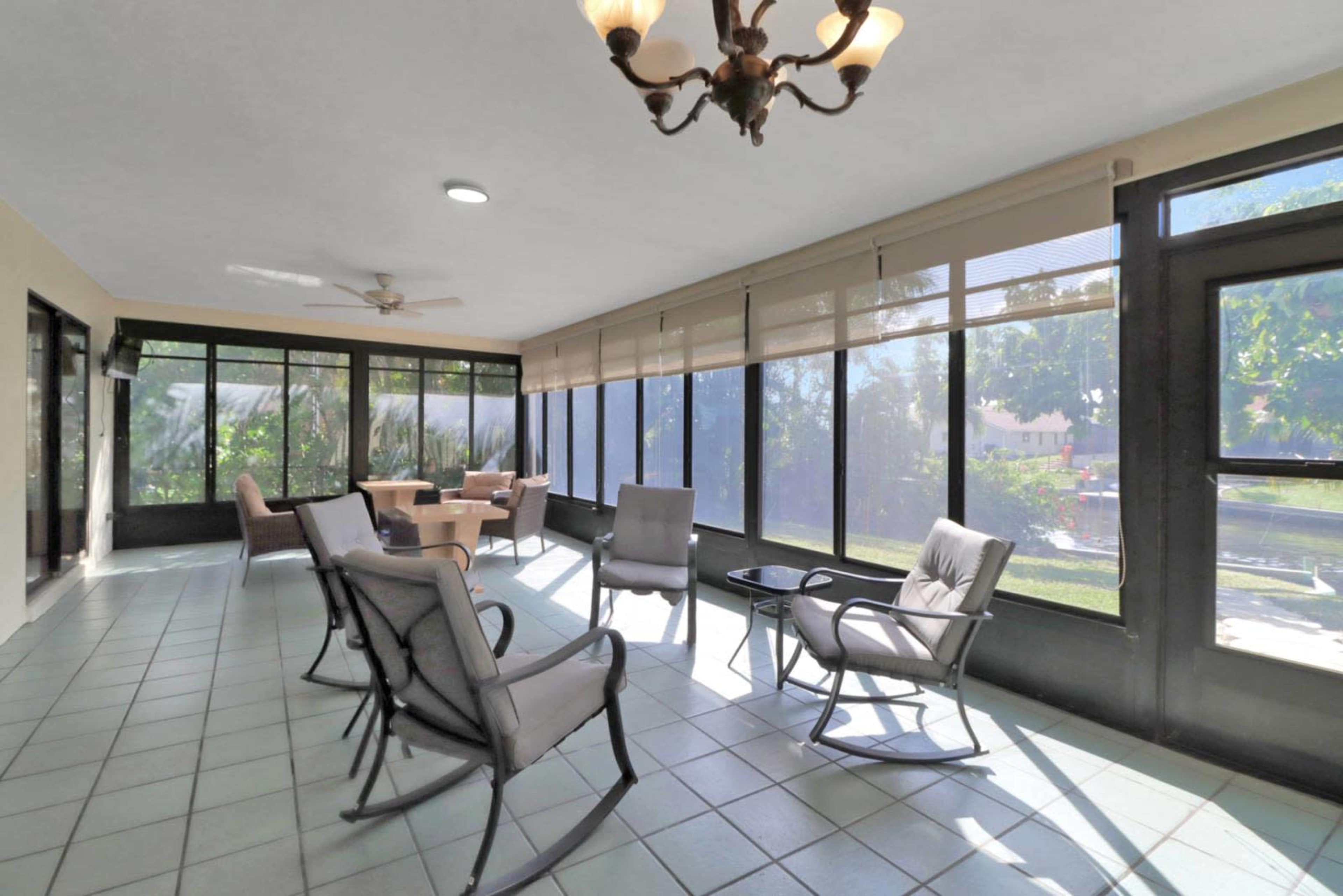 The image shows a sunlit screened porch with several chairs, a small table, and large windows displaying a view of greenery outside.