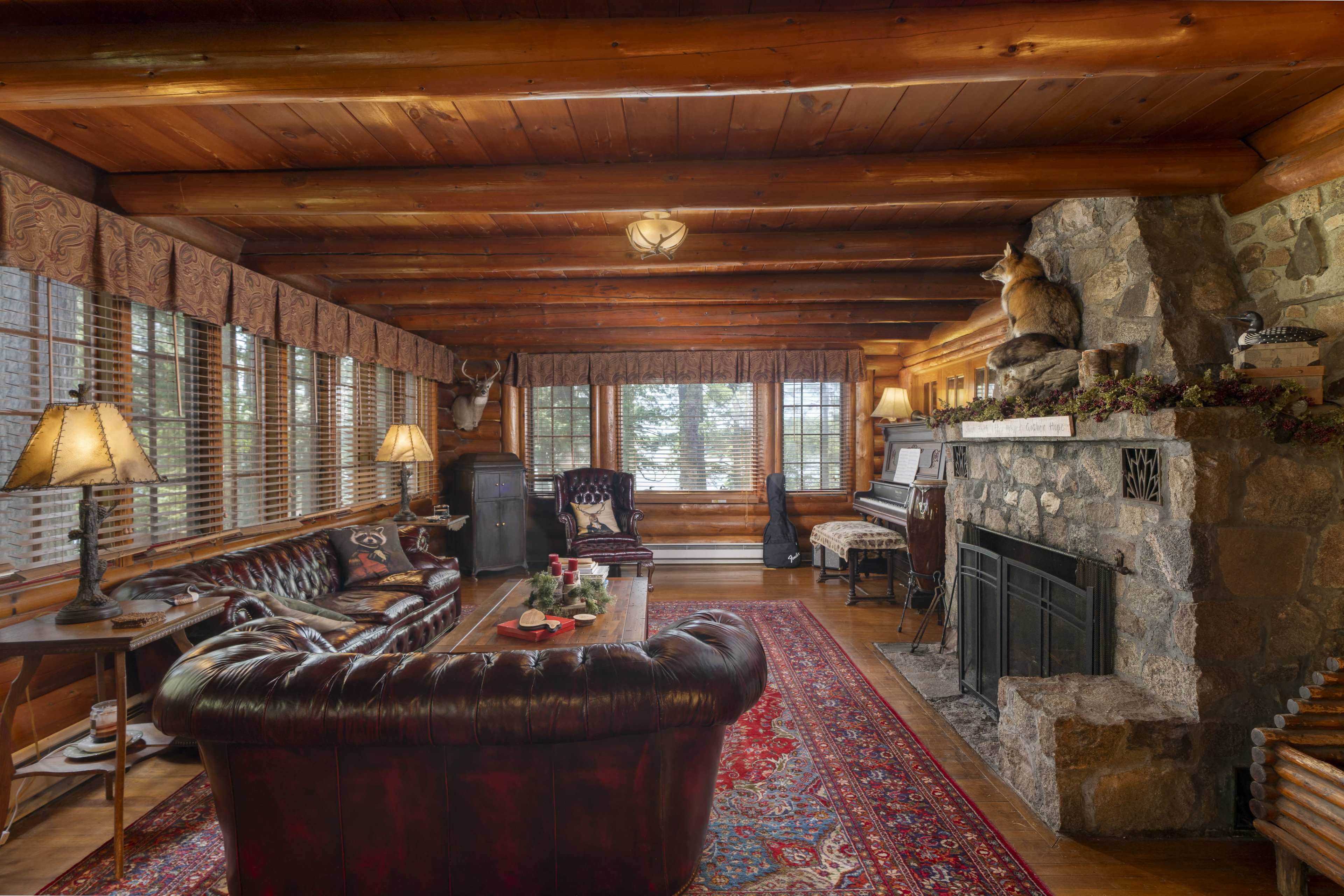 The image shows a cozy living room with wooden beams, a stone fireplace, a dark leather couch, and large windows adorned with patterned curtains.
