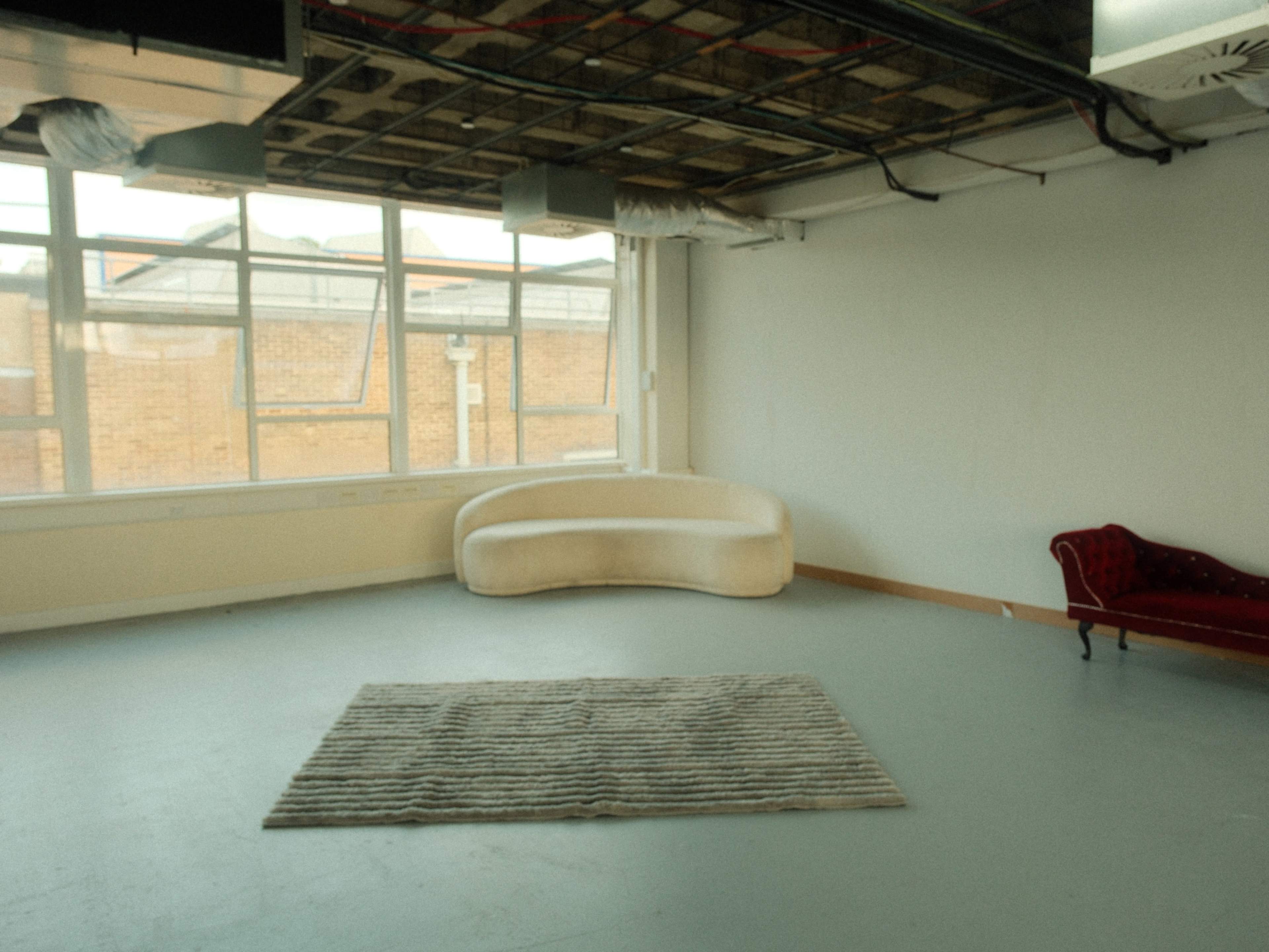 The image shows a spacious, empty room with a large window, a curved white couch, a red sofa, and a striped rug on the floor.