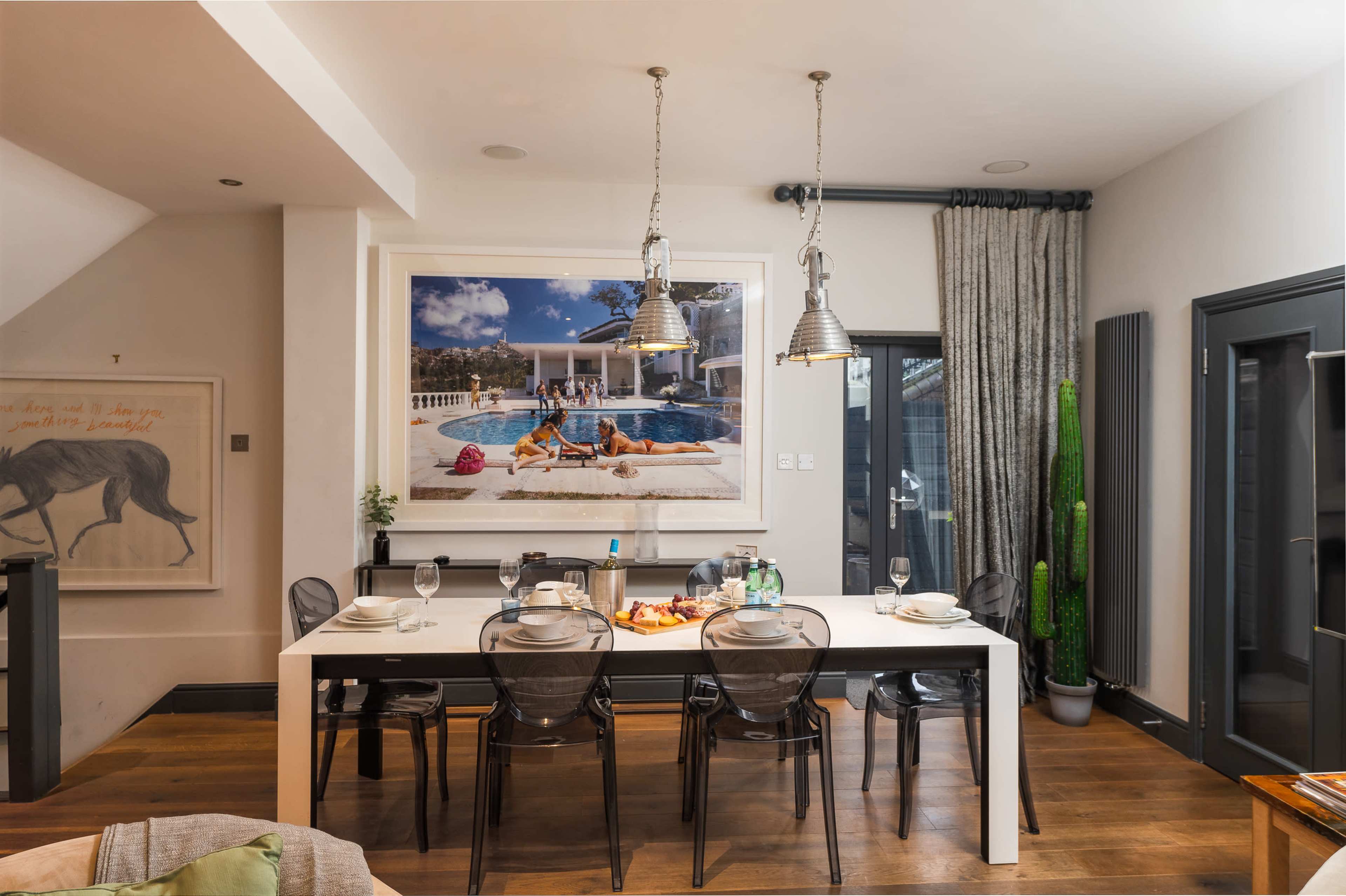 The image shows a modern dining area with a long table set for a meal, featuring a large photograph of a pool scene on the wall.