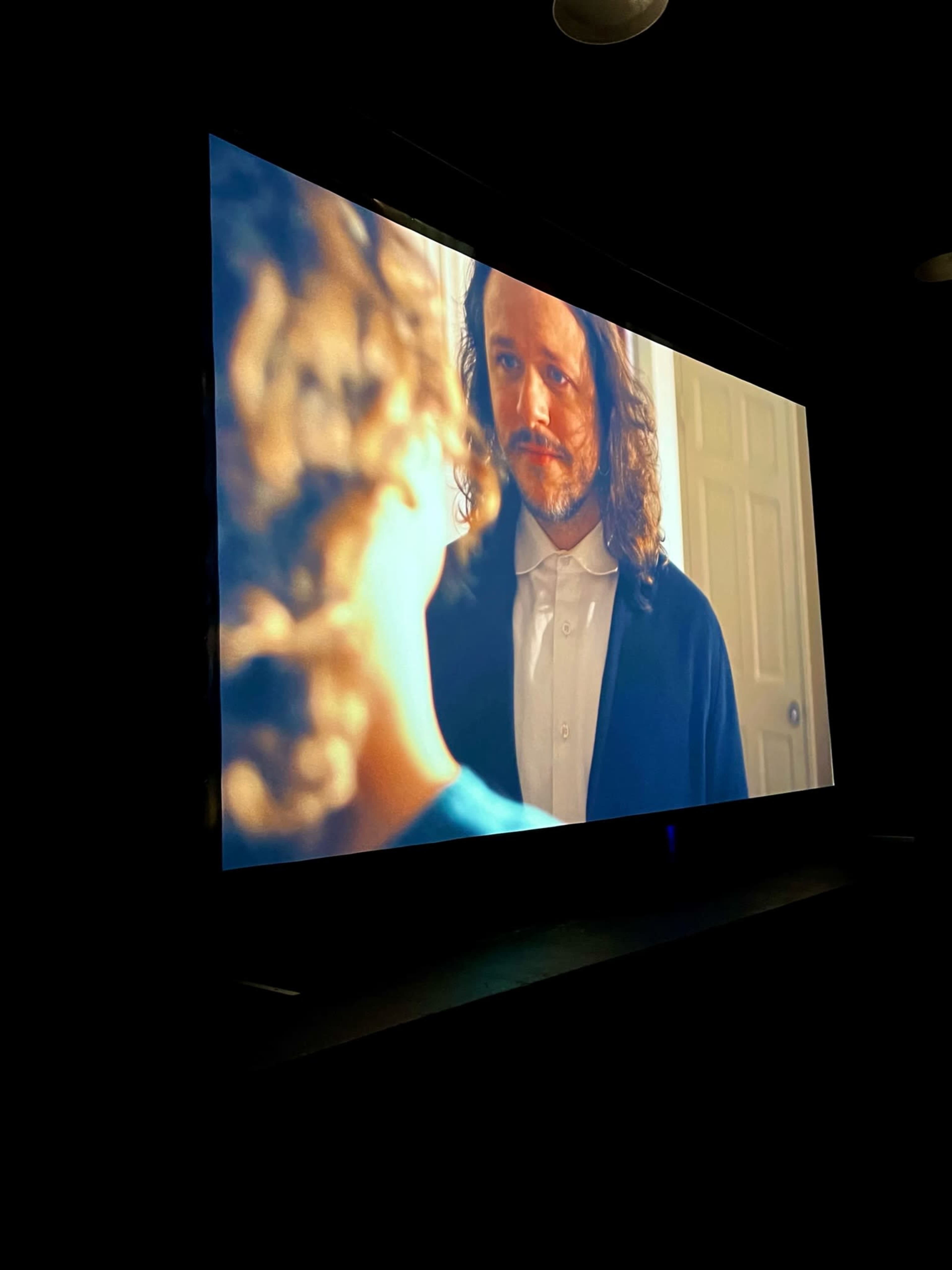 A projection of a man with long hair and a beard speaks to a child in a dimly lit room.