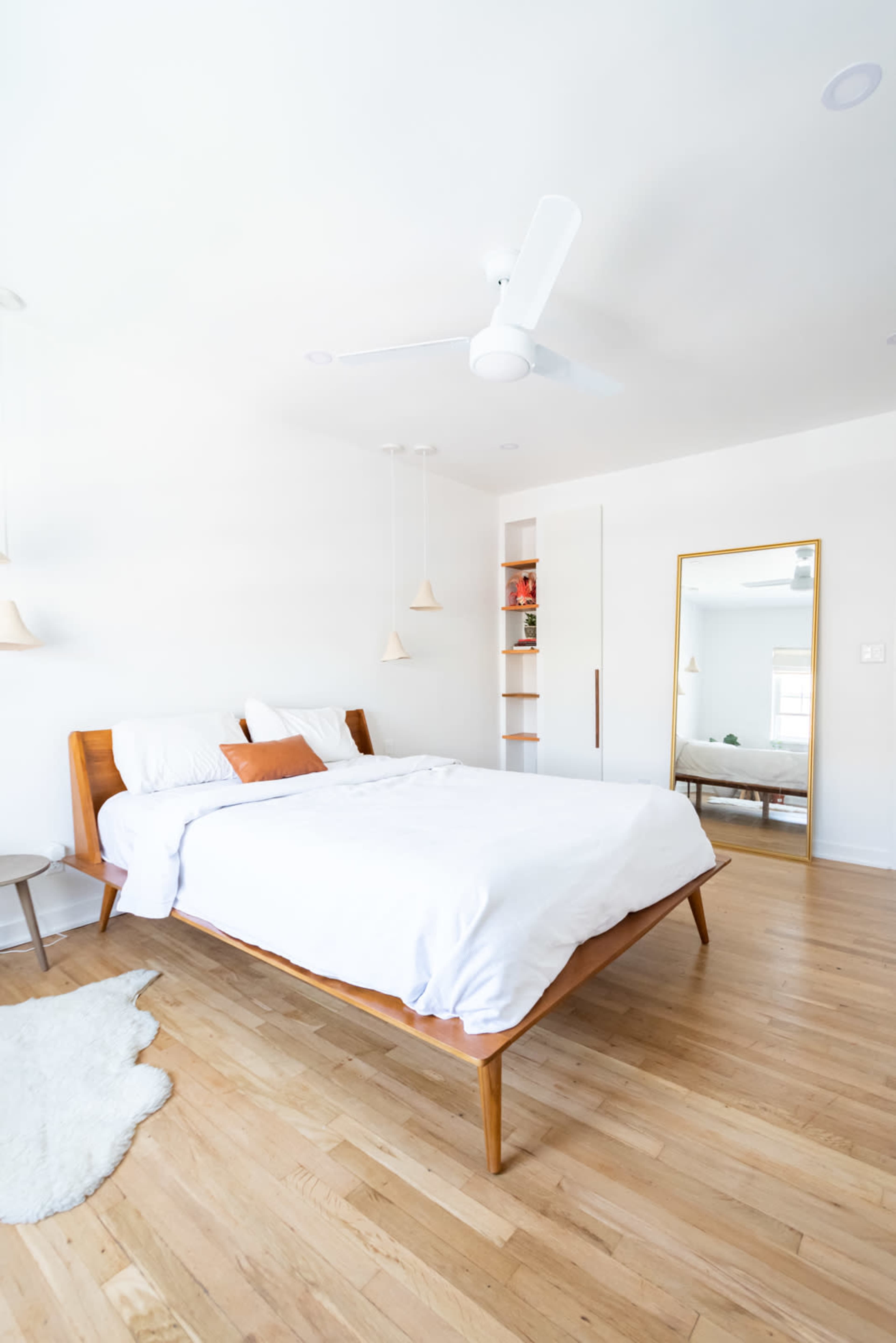 A light-filled bedroom features a wooden bed with a white comforter, a small side table, and a large mirror reflecting a shelf with neatly arranged items.