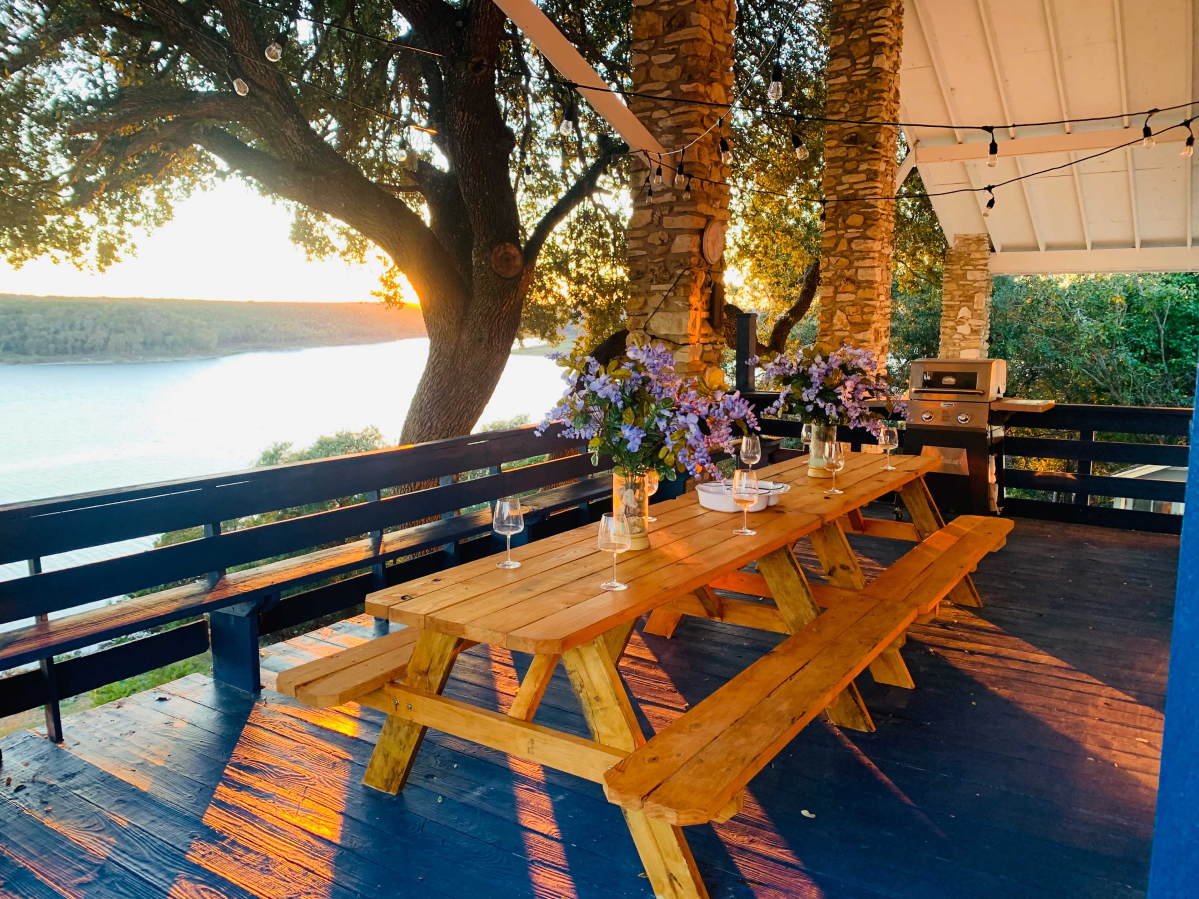 A wooden picnic table set for a meal on a deck overlooking a lake, with flowers and glasses arranged neatly on the tables and a sunset in the background.