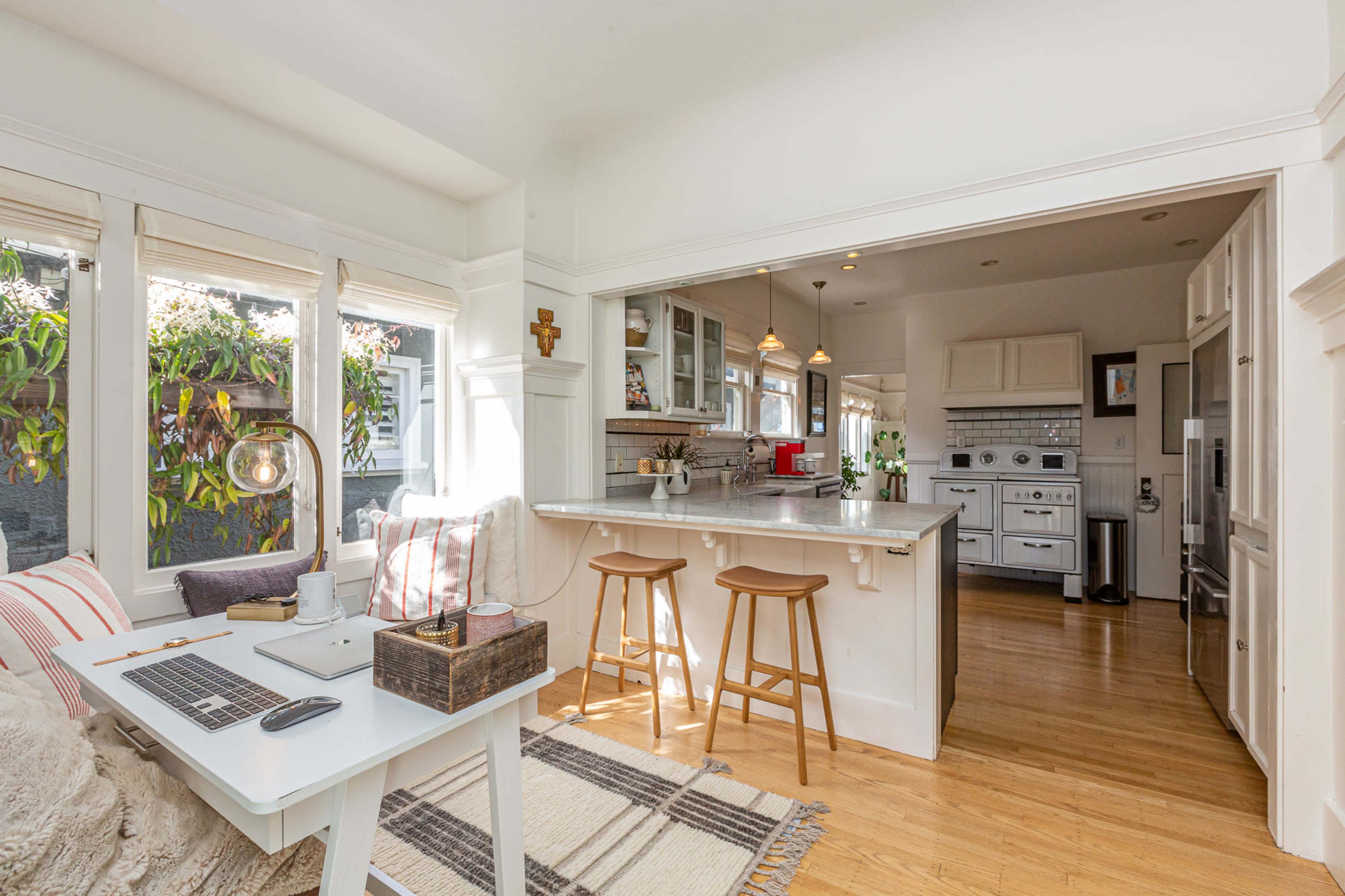 A bright kitchen with a minimalistic dining area featuring two wooden stools, a white table, and large windows allowing natural light to enter.