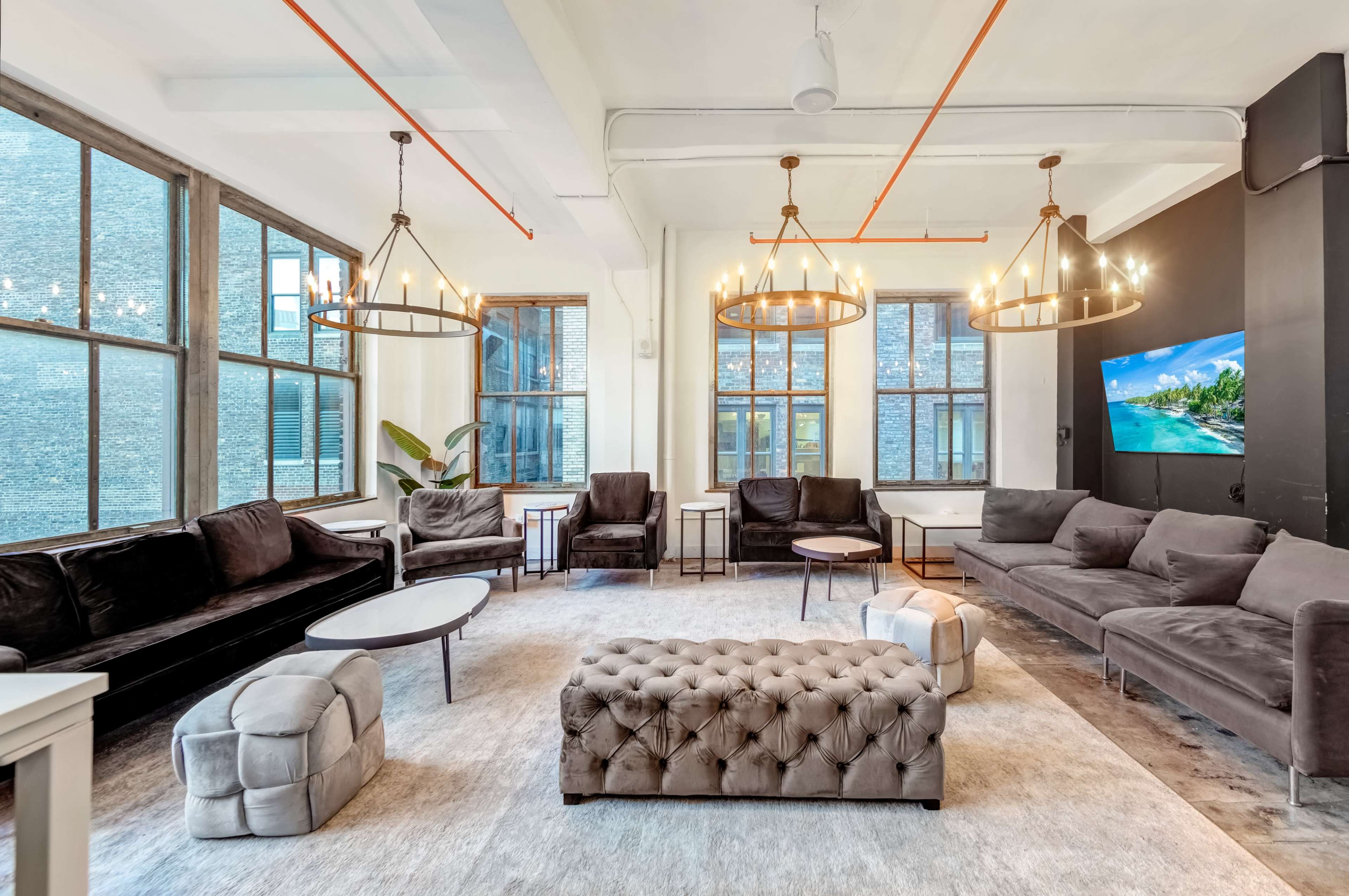 A modern lounge area with large windows, several seating arrangements, a tufted ottoman, and decorative lighting fixtures.