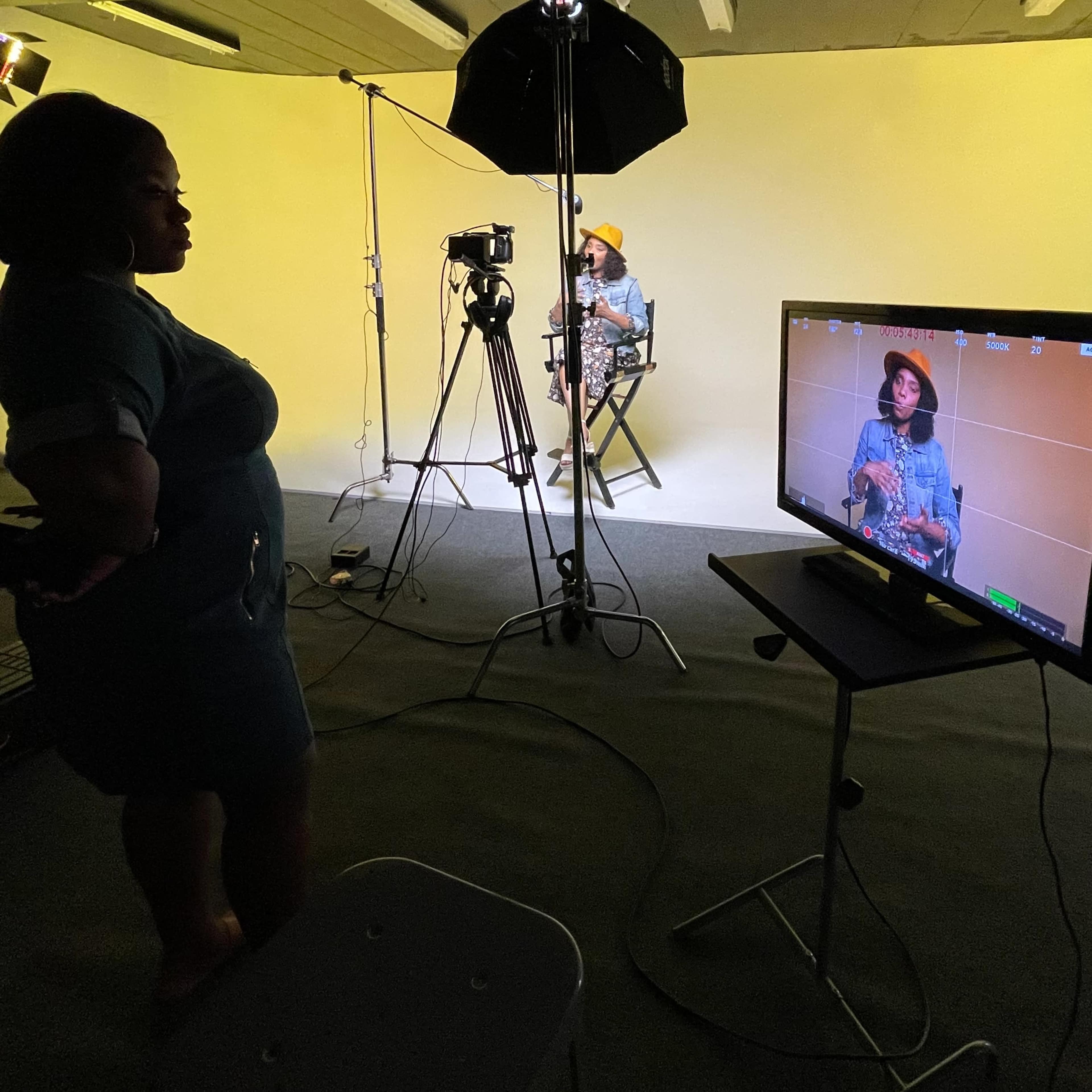 A woman stands in front of a seated individual who is being filmed against a plain yellow backdrop, with studio lighting and a camera monitor visible.