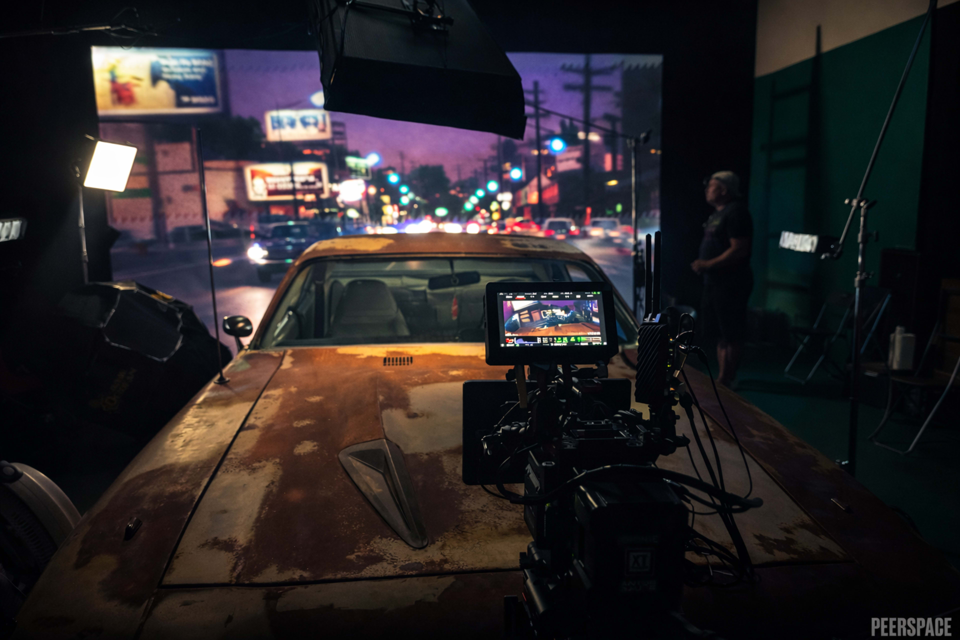 A weathered car is positioned in front of a backdrop featuring a city street scene, with lights and camera equipment set up for a film shoot.