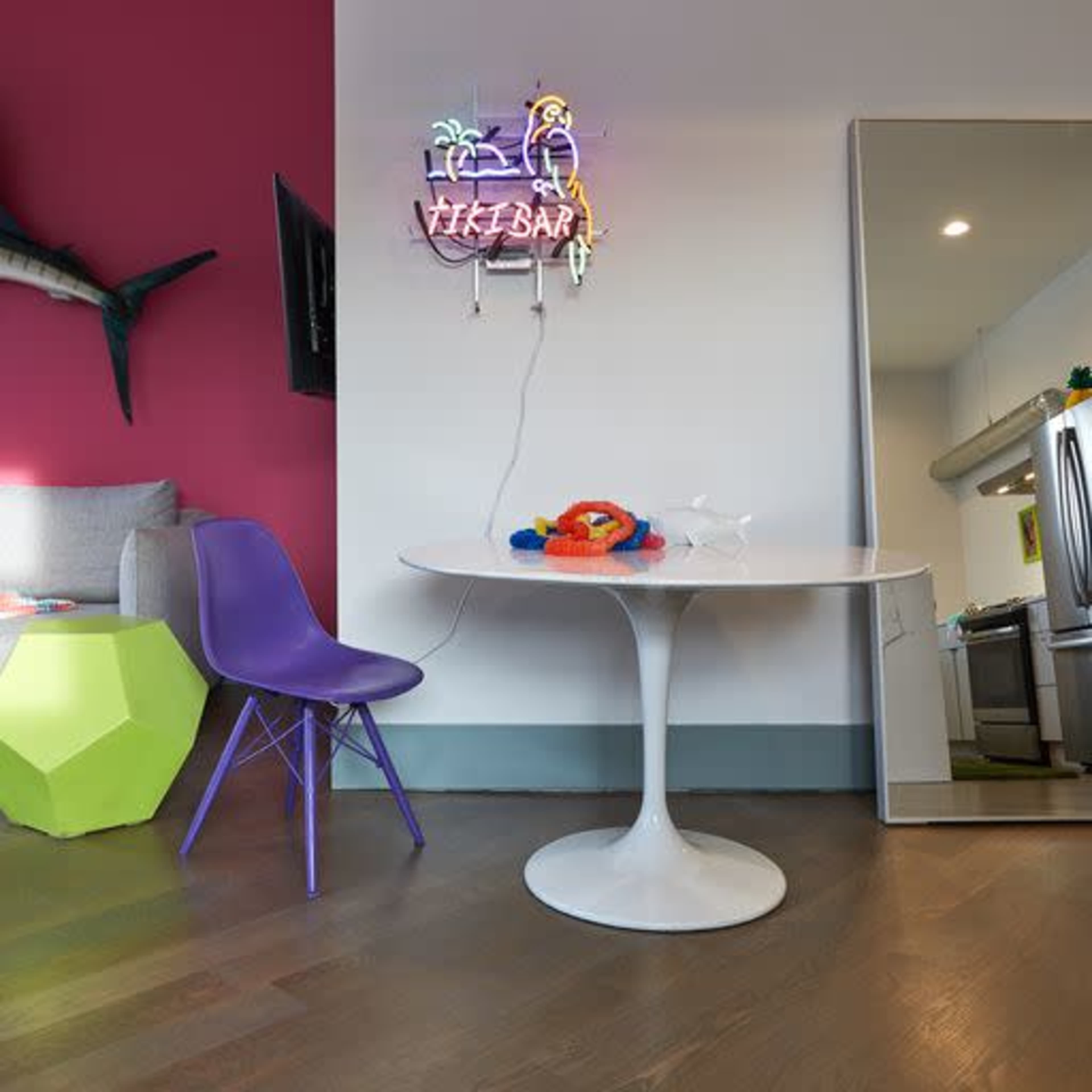 The image features a modern living space with a round white table, colorful chair, and a neon "Tiki Bar" sign against a vibrant pink wall.
