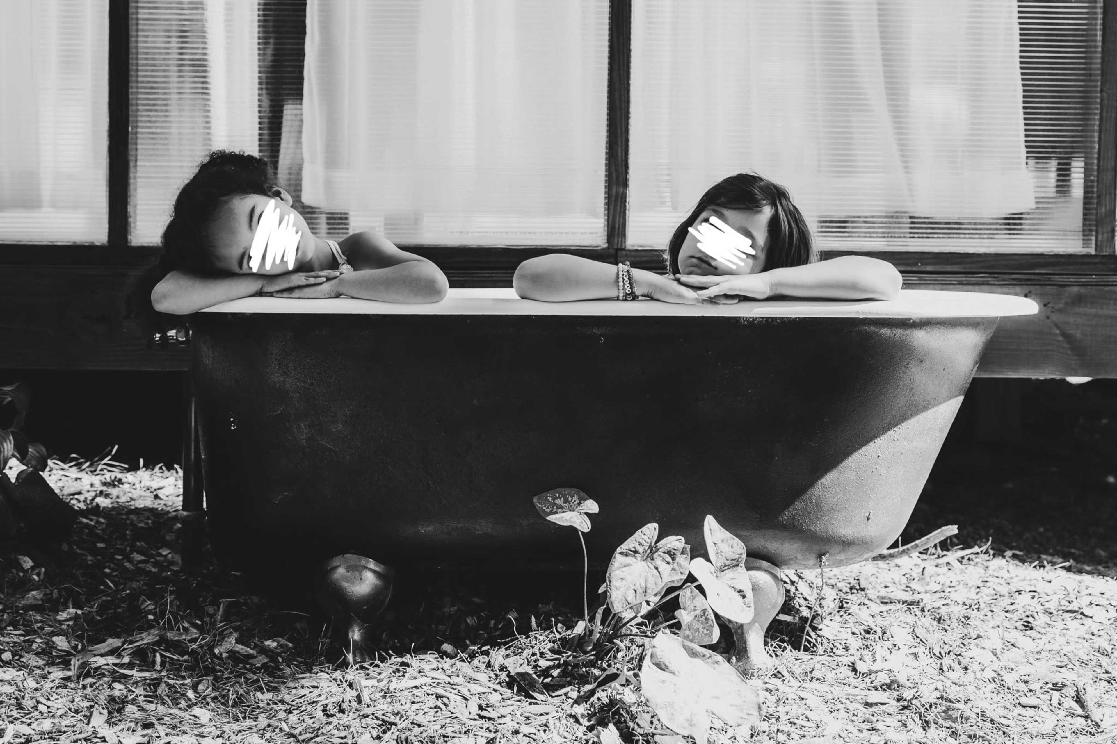 Two women relax in a vintage bathtub surrounded by plants and soft natural light.