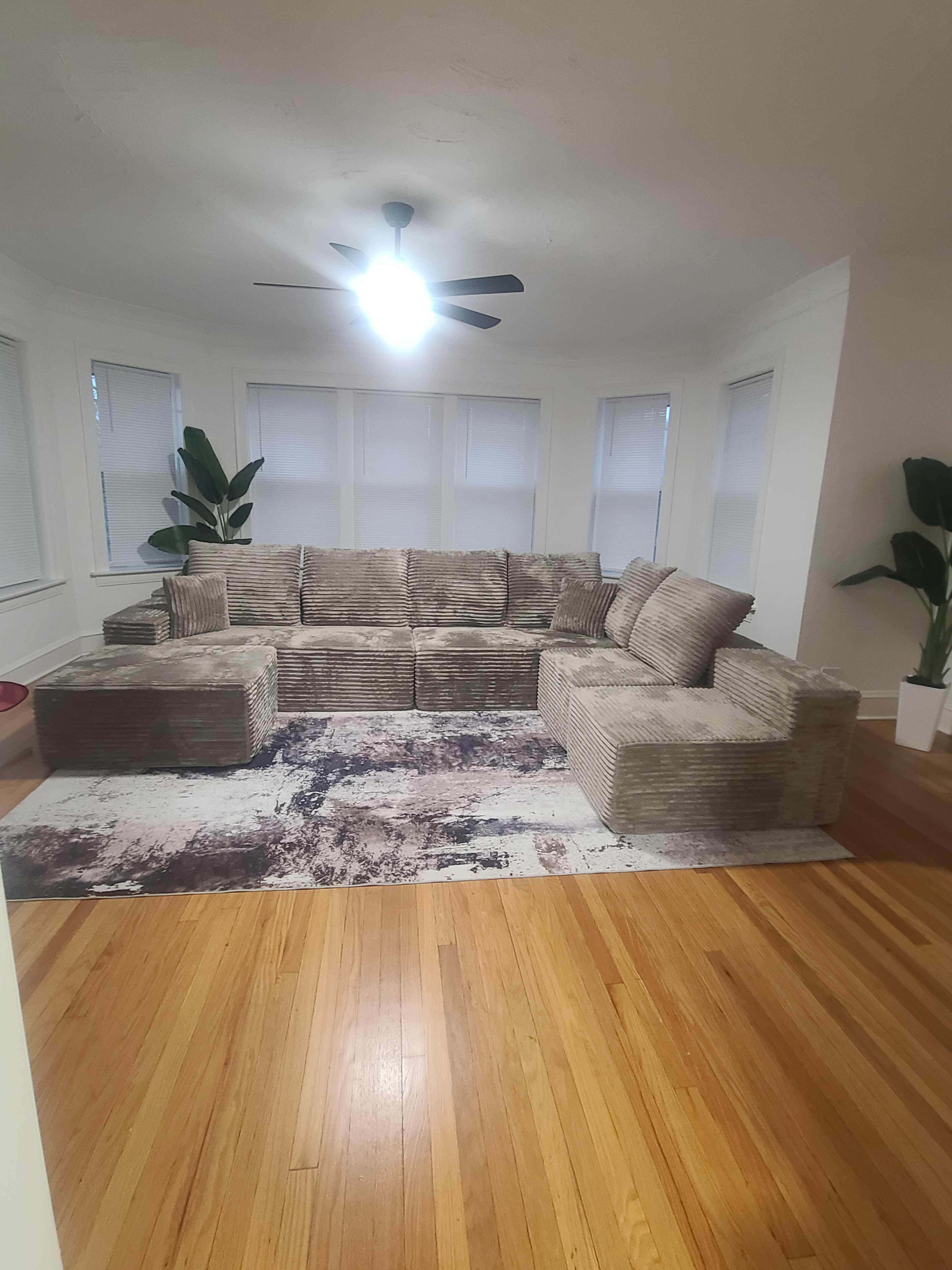 The image shows a large, light-colored sectional sofa arranged in a bright room with hardwood floors, a ceiling fan, and potted plants.