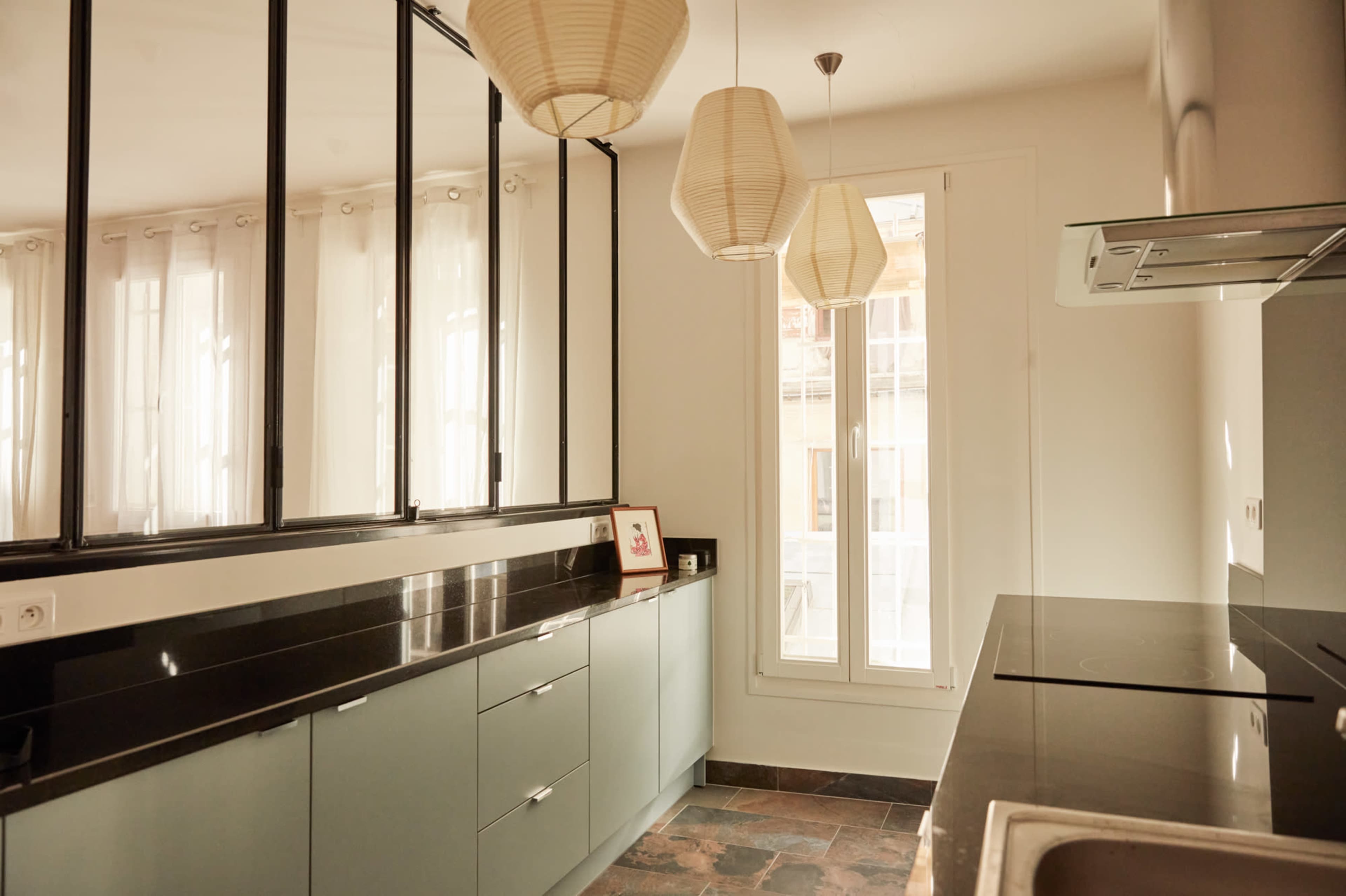 A modern kitchen featuring light green cabinets, a black countertop, and large windows with sheer curtains.