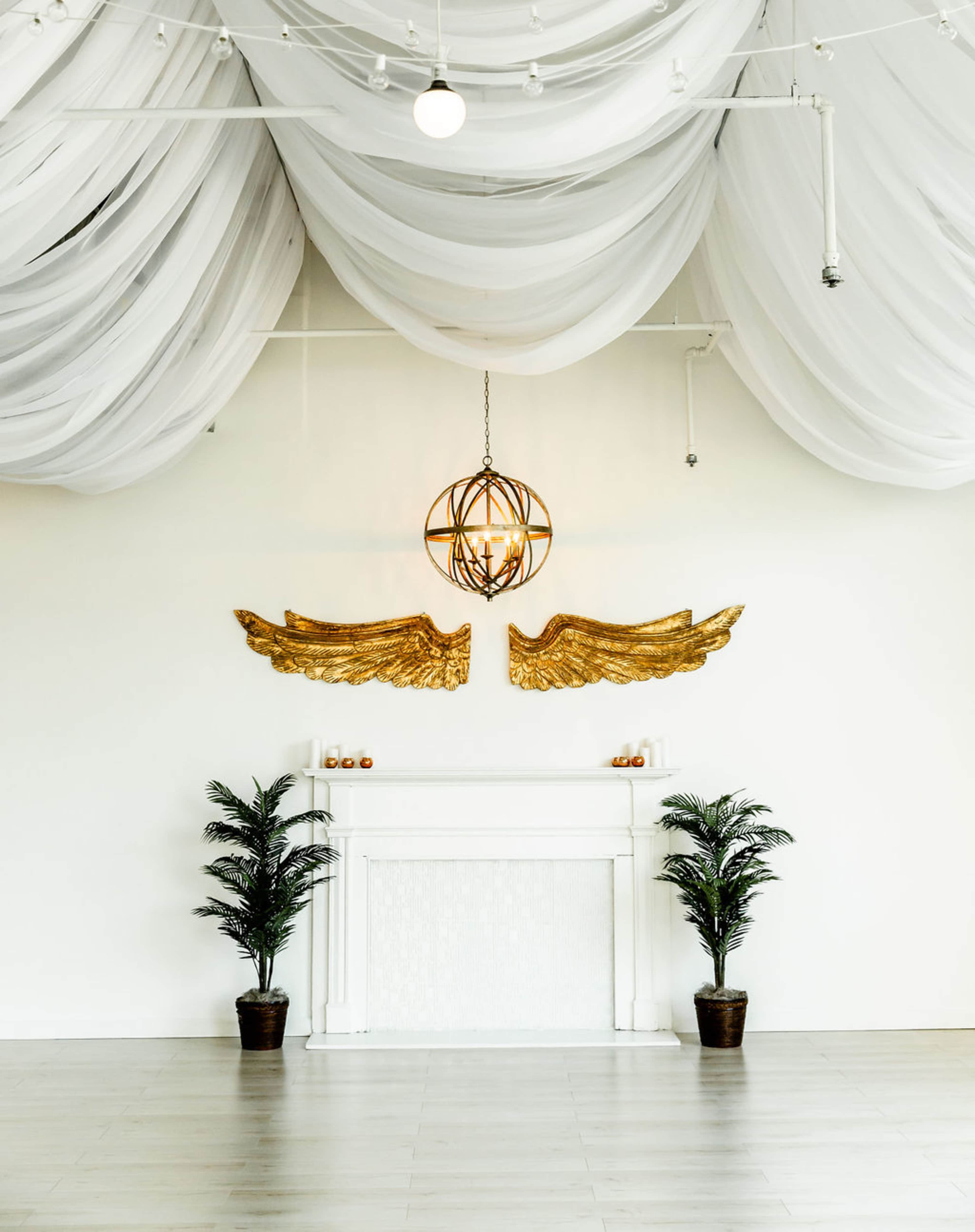 A spacious room with white draped fabric hanging from the ceiling, a golden winged decoration on the wall, and a white mantel flanked by two potted plants.